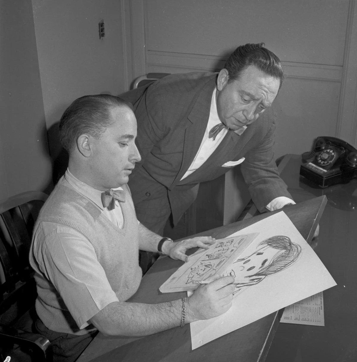 Irwin Hasen (seated at his drawing table) and Gus Edson (standing) discuss Dondi.