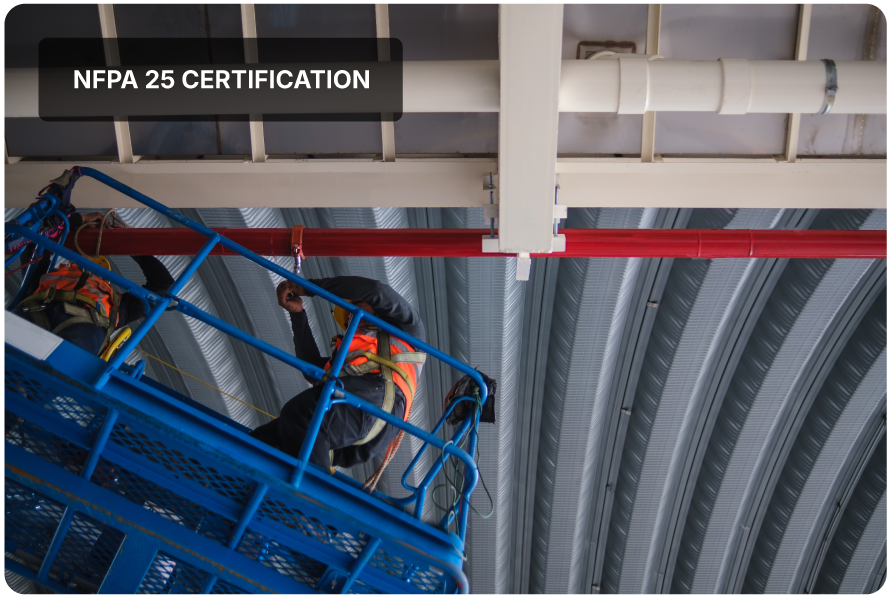 Worker wearing safety harness on an elevated blue lift inside an industrial building with metal roof and red pipe overhead.