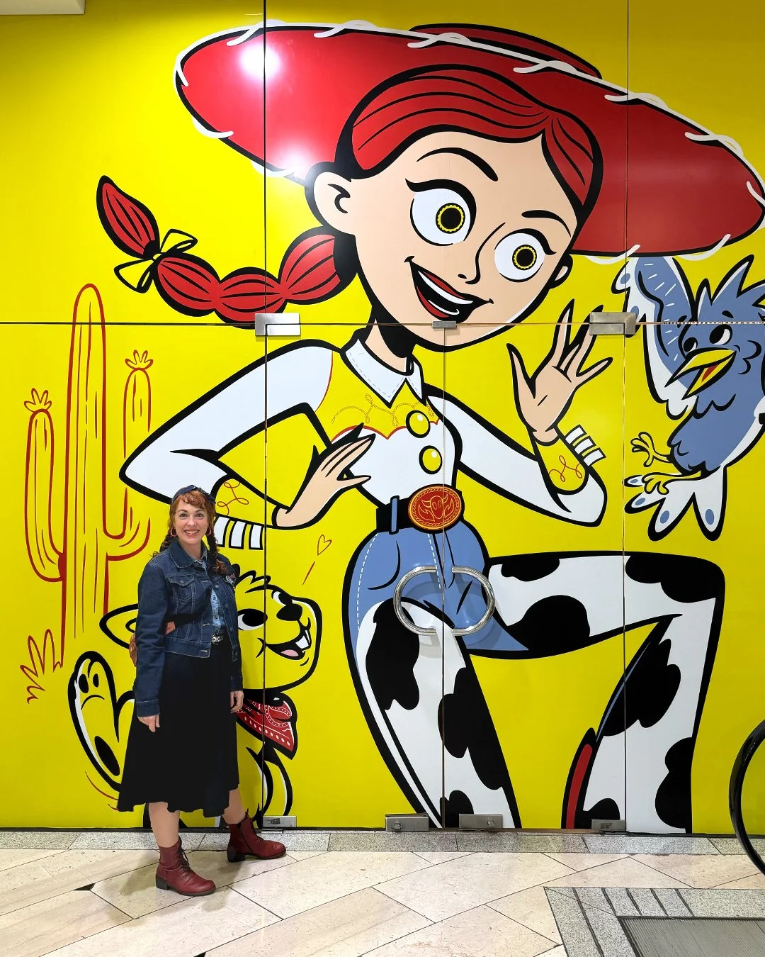 A woman standing in front of a large colorful mural of Jessie from Toy Story with her hand raised, a blue bird, a cowgirl cat, and a cactus in the background.