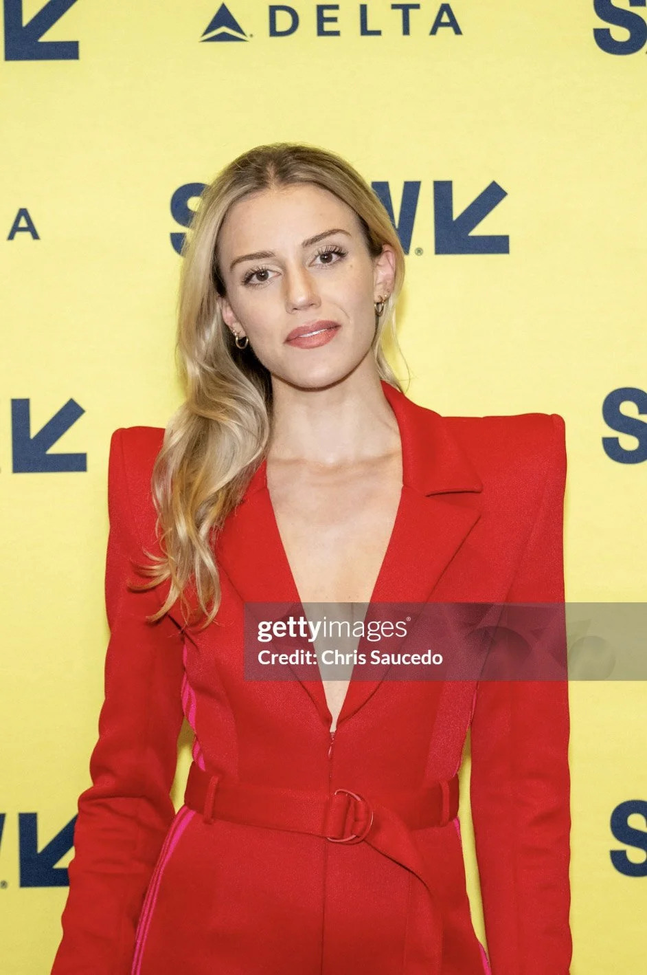 Katherine Cozumel on the red carpet at SXSW (South by Southwest), she has long blonde hair and is wearing a red suit with a deep neckline, standing in front of a yellow backdrop with blue text and SXSW logos.