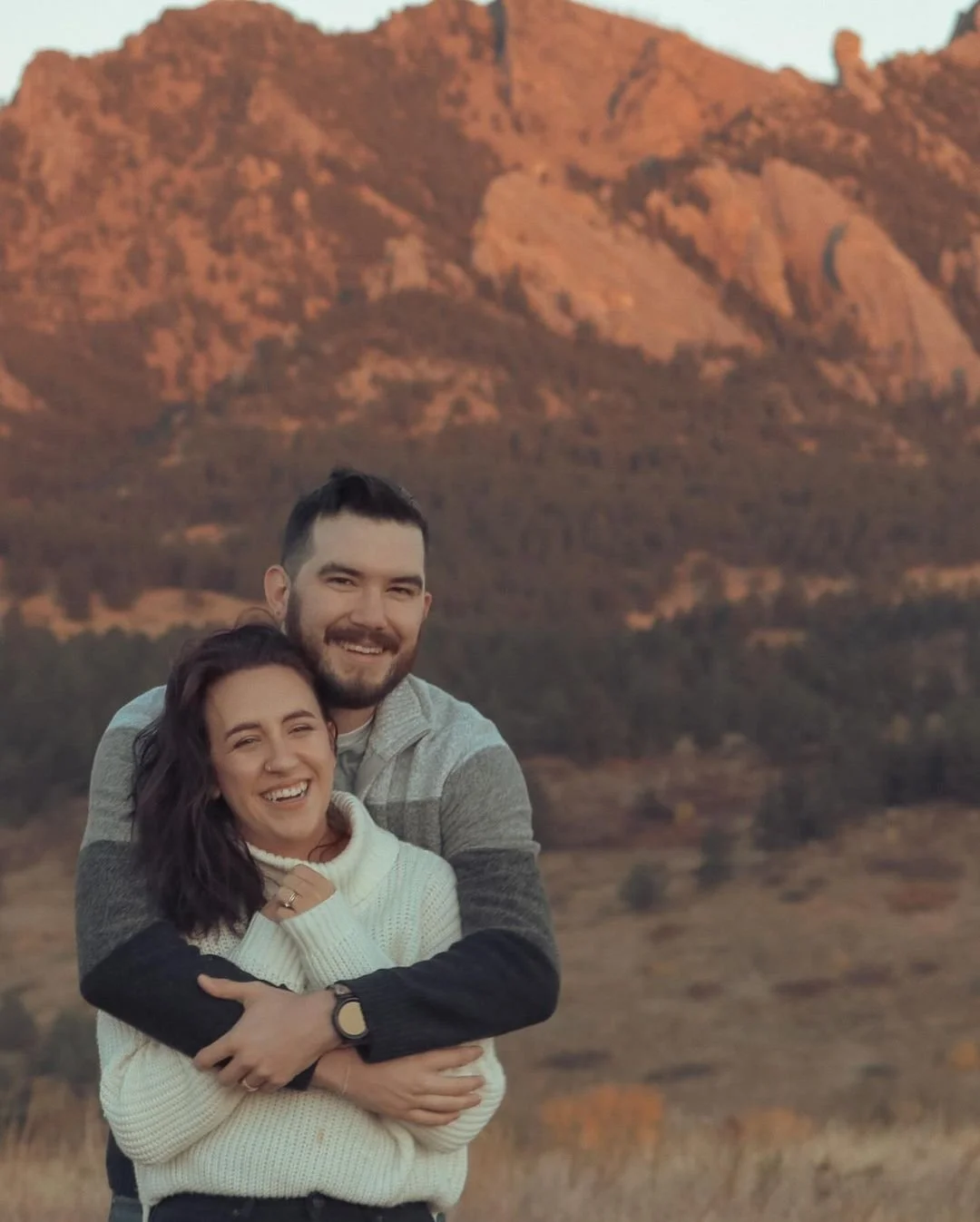 A smiling couple embracing outdoors with a backdrop of mountains at sunset.