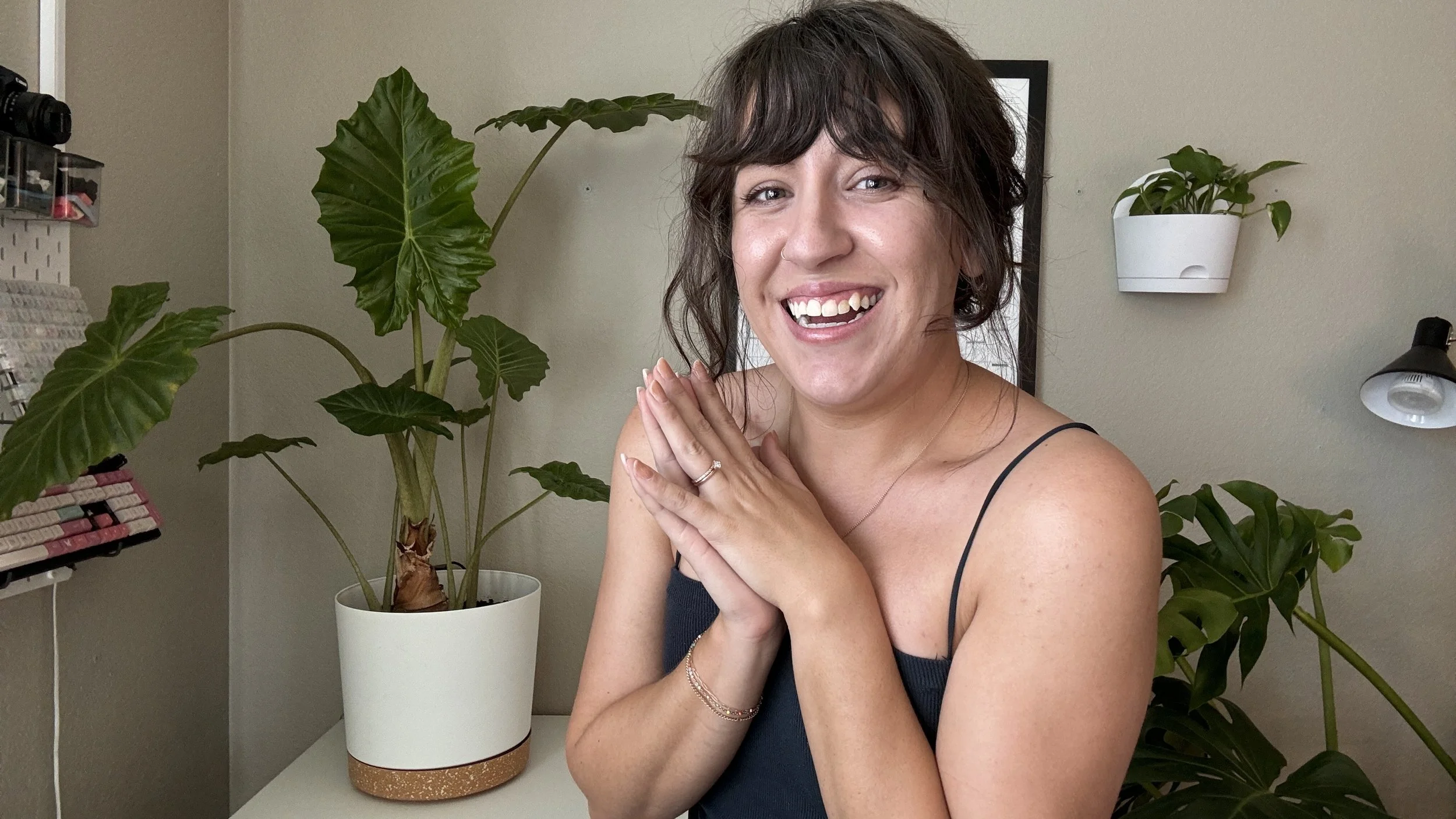 A woman with dark wavy hair, wearing a black spaghetti strap top, smiling with her hands clasped near her face, surrounded by green houseplants indoors.