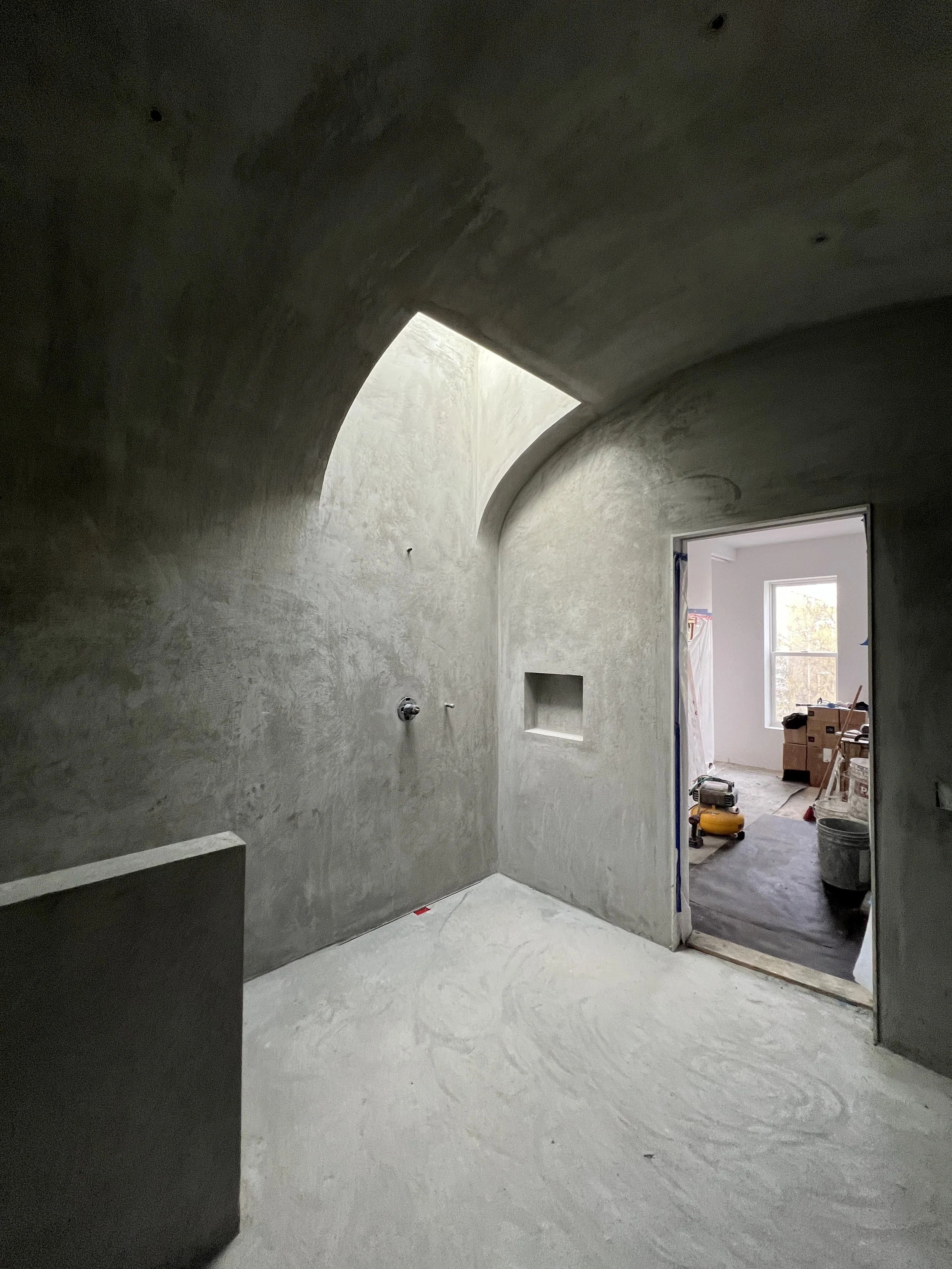 Interior of a room under construction with curved walls and a skylight, partially finished floor, and an open doorway leading to a brighter room.