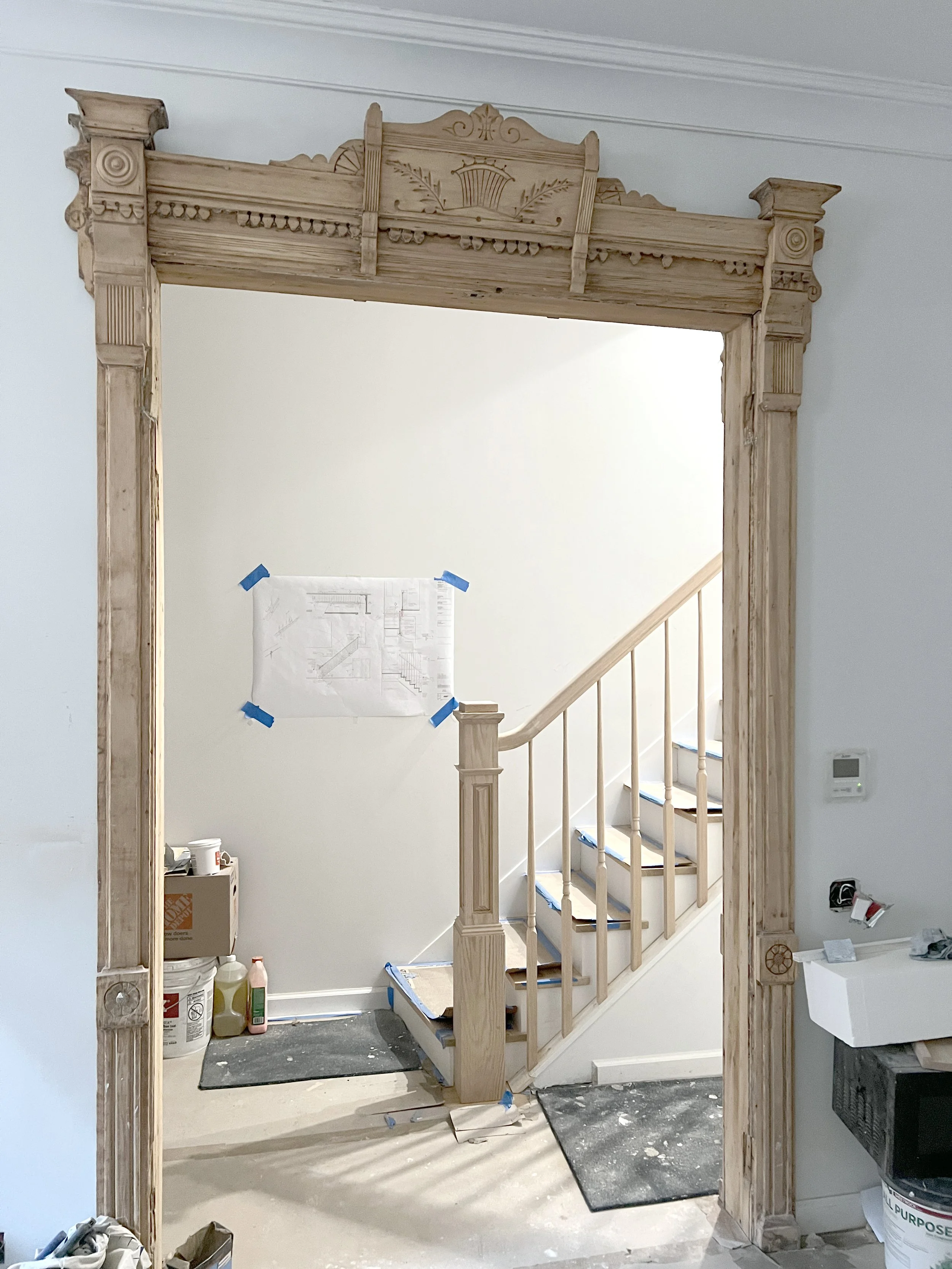 Wooden doorway frame under construction with detailed carvings. Staircase with wooden railing visible through the opening, and construction items like paint cans, brushes, and cardboard boxes on the floor.