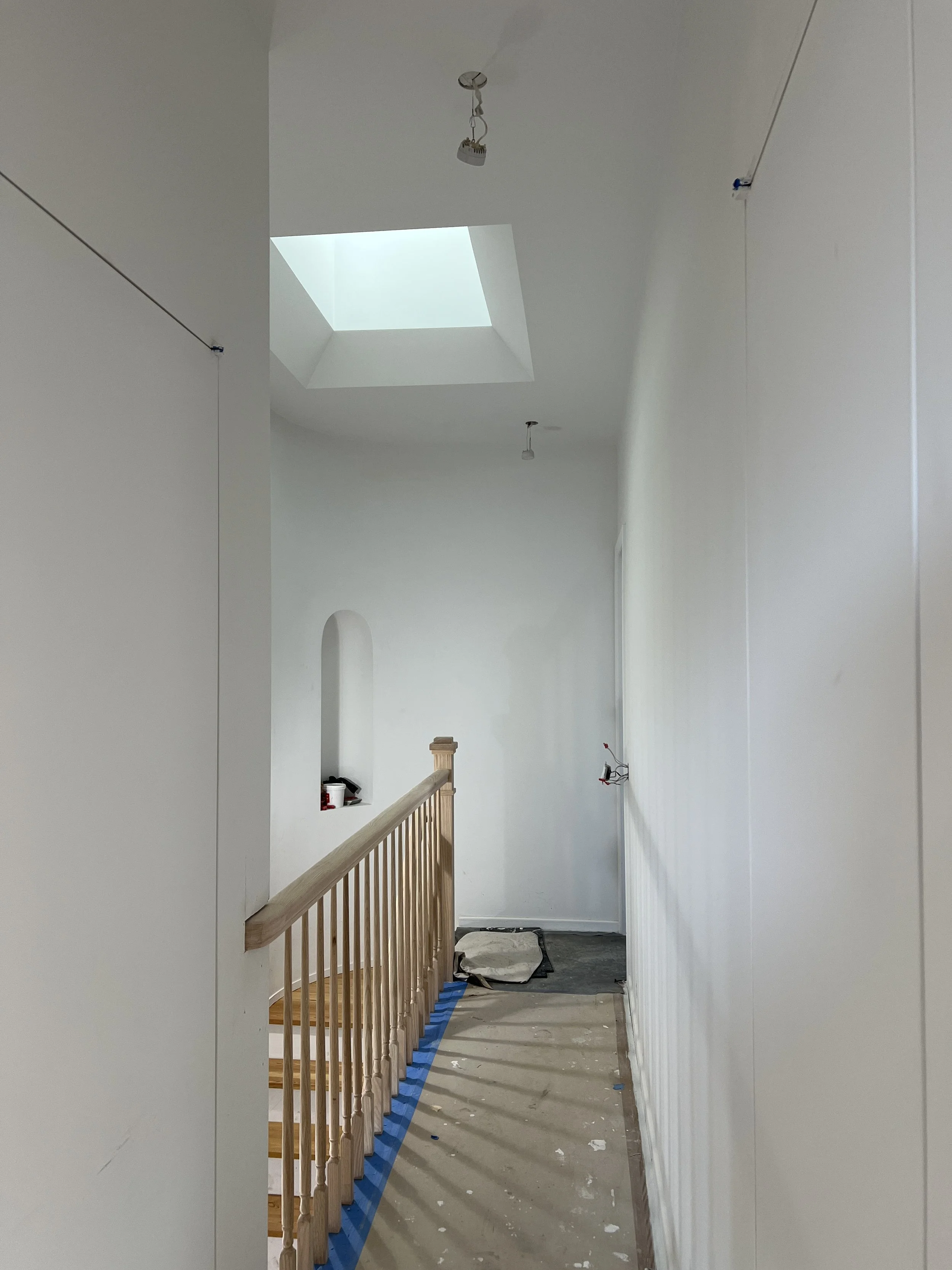 Interior of a house under construction with white walls, a skylight, and a wooden railing along a staircase opening, exposed electrical wiring, and construction materials on the floor.