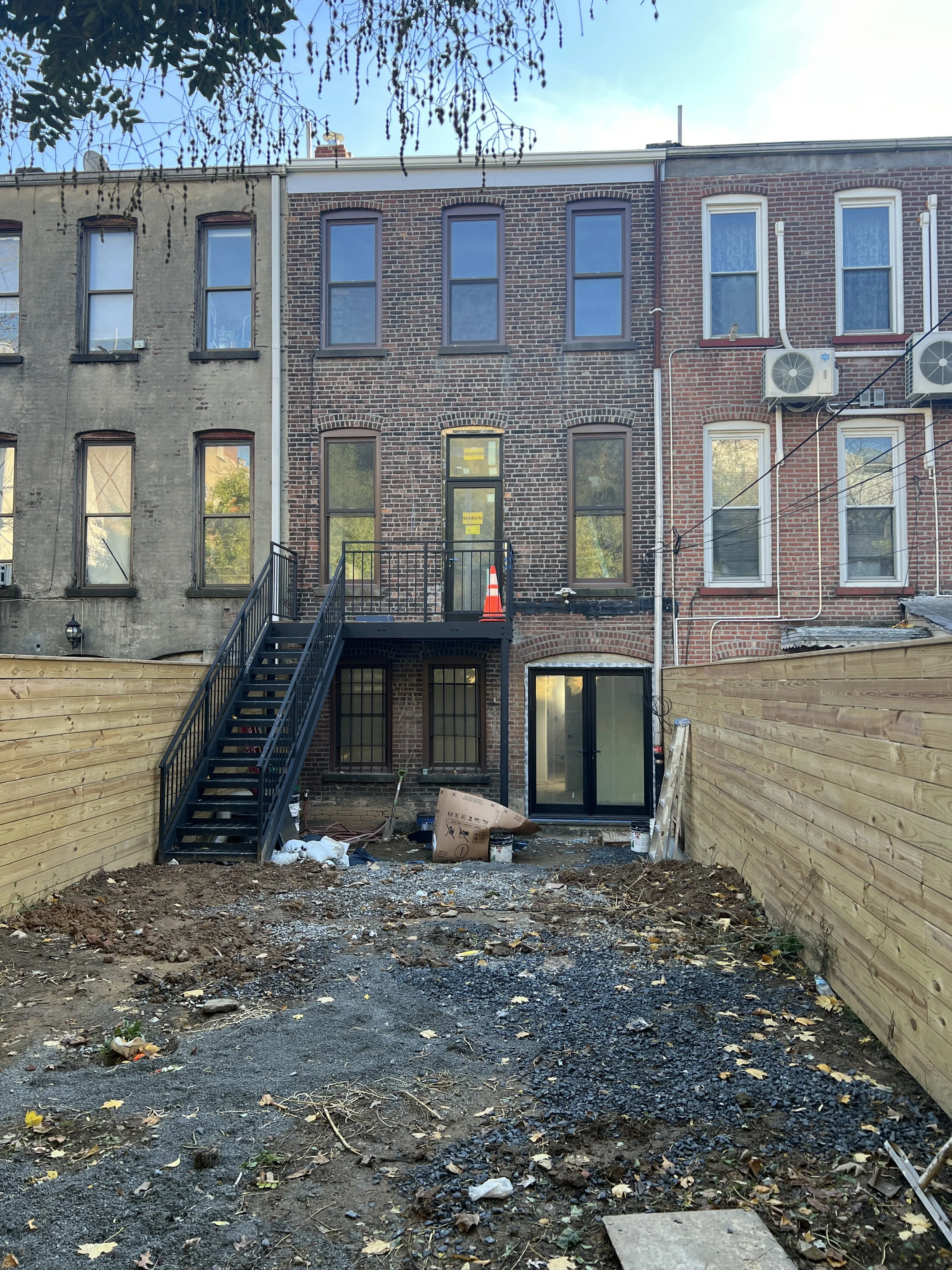 Backyard under construction of a row house with a wooden fence, gravel, and building materials, with an exterior staircase leading to the upper level.