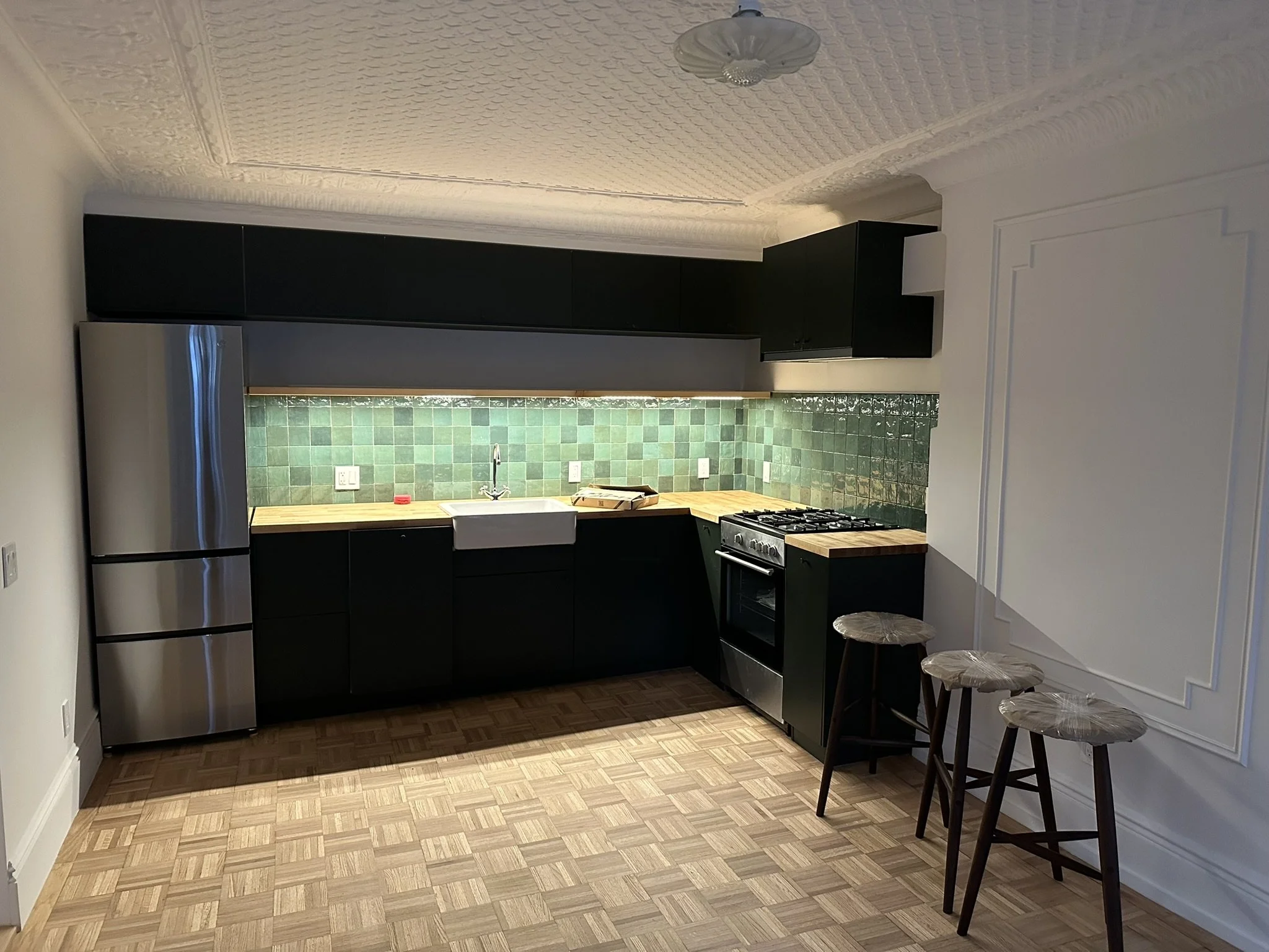 Kitchen with black cabinets, green tiled backsplash, white farmhouse sink, silver refrigerator, and wooden kitchen stools