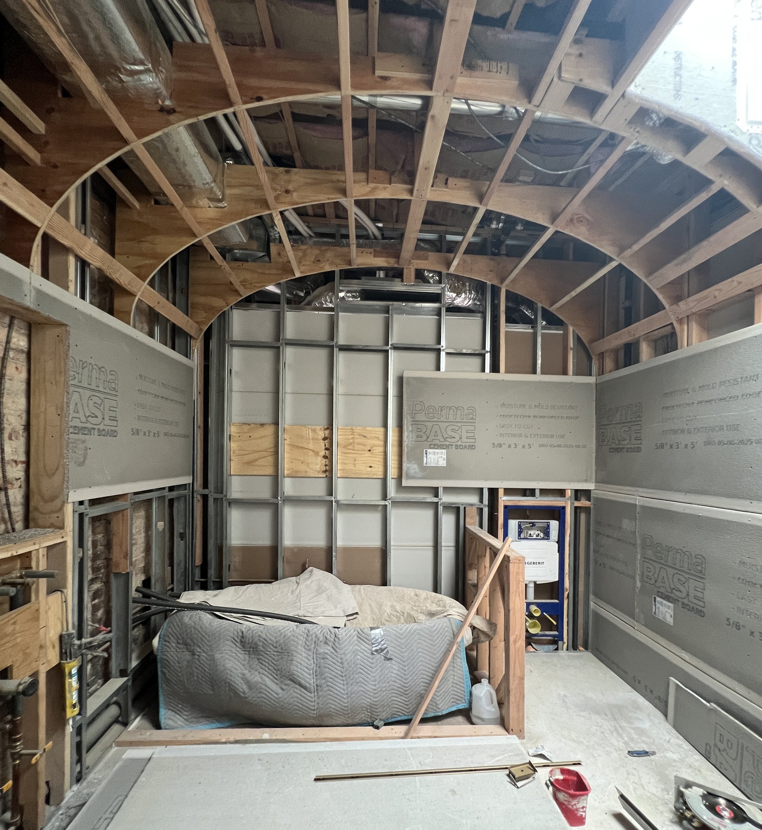 View of a room under construction with exposed framing, insulation, and plumbing. A bathtub covered with a tarp and a water bottle inside a framed enclosure are visible. Construction tools and materials are scattered on the floor.