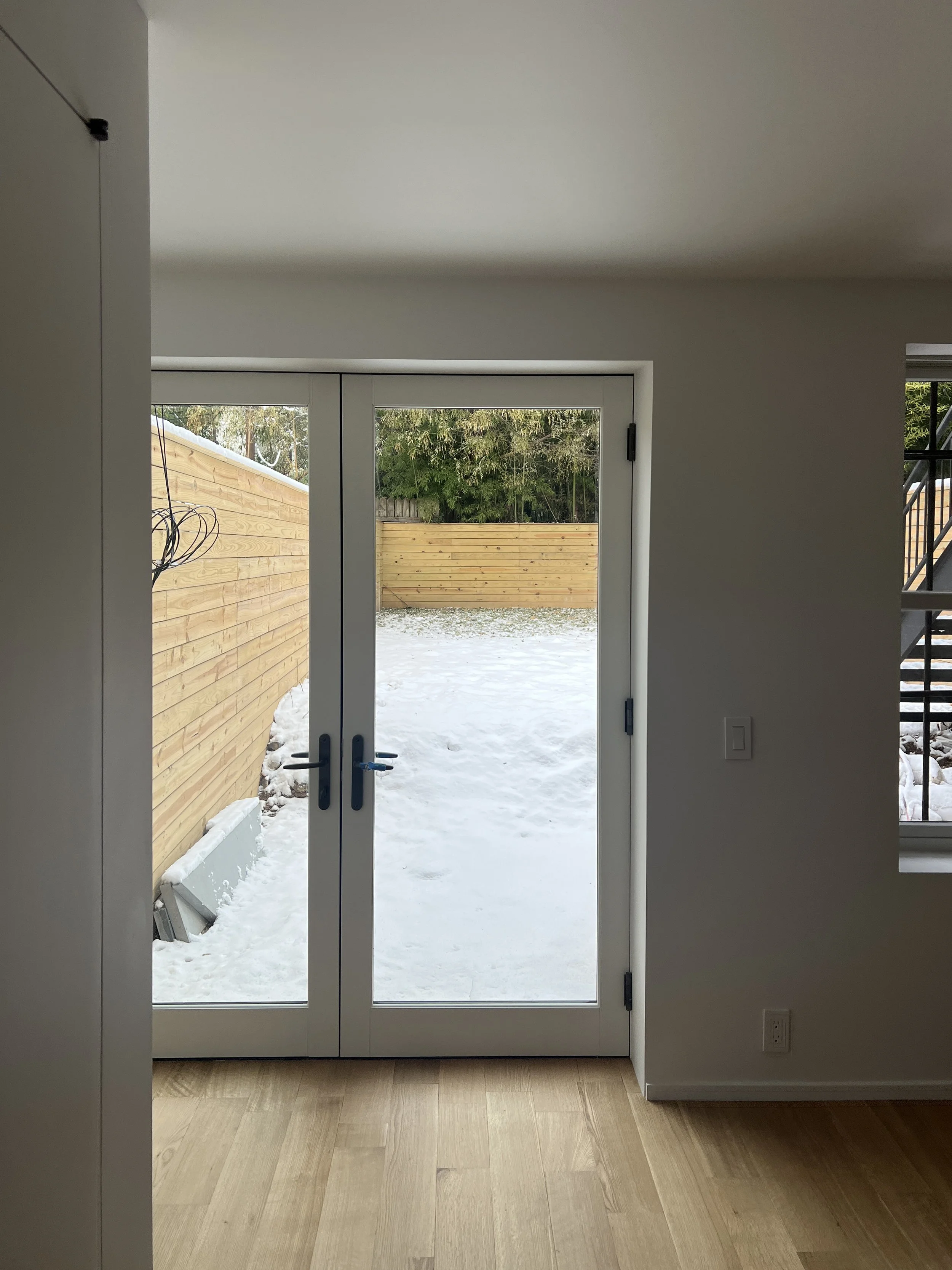 Interior view of a room with wooden floor, white walls, and glass double doors leading to a snow-covered backyard with a wooden fence.