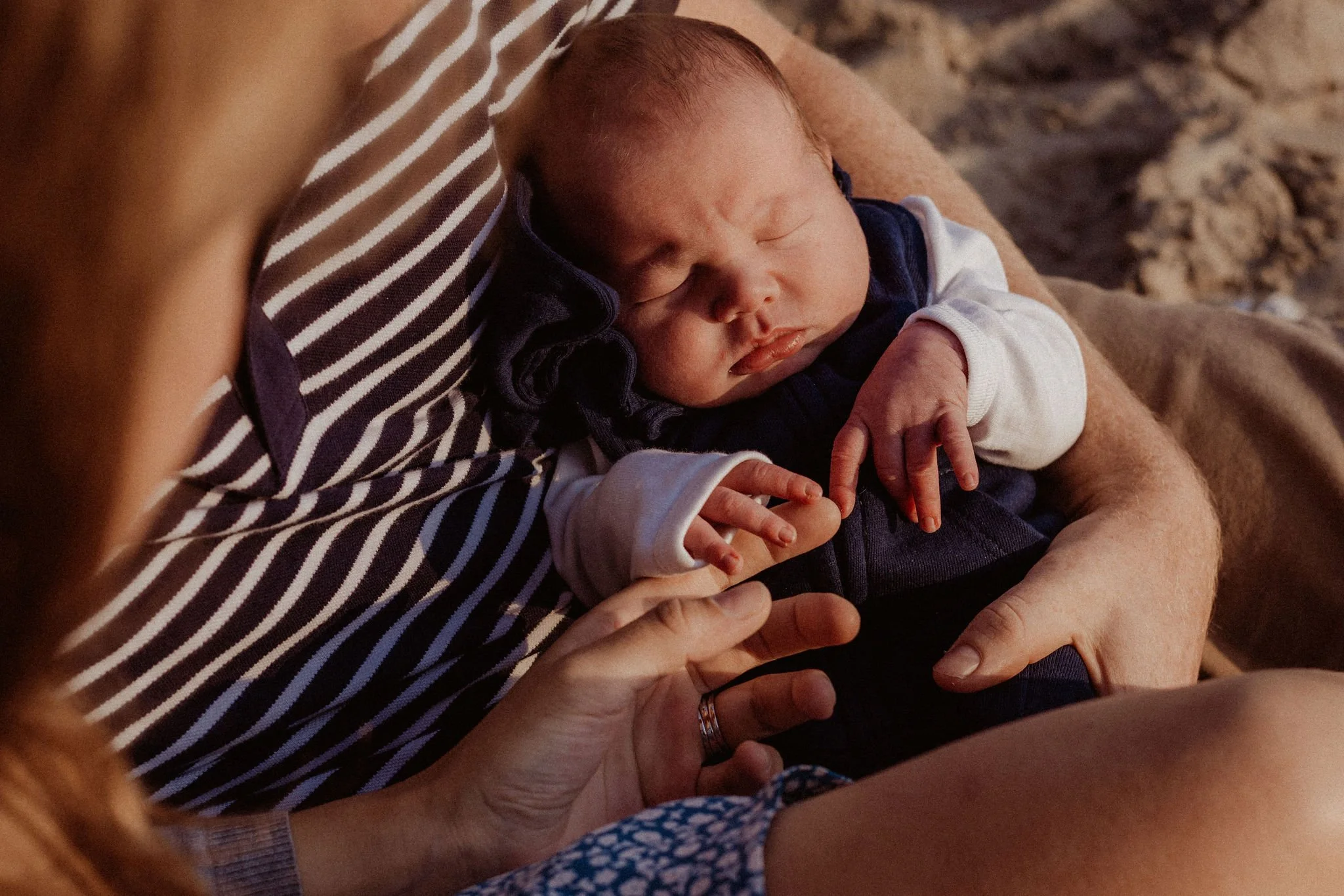 A sleeping baby held close by a parent, on a beach with sandy ground in the background.