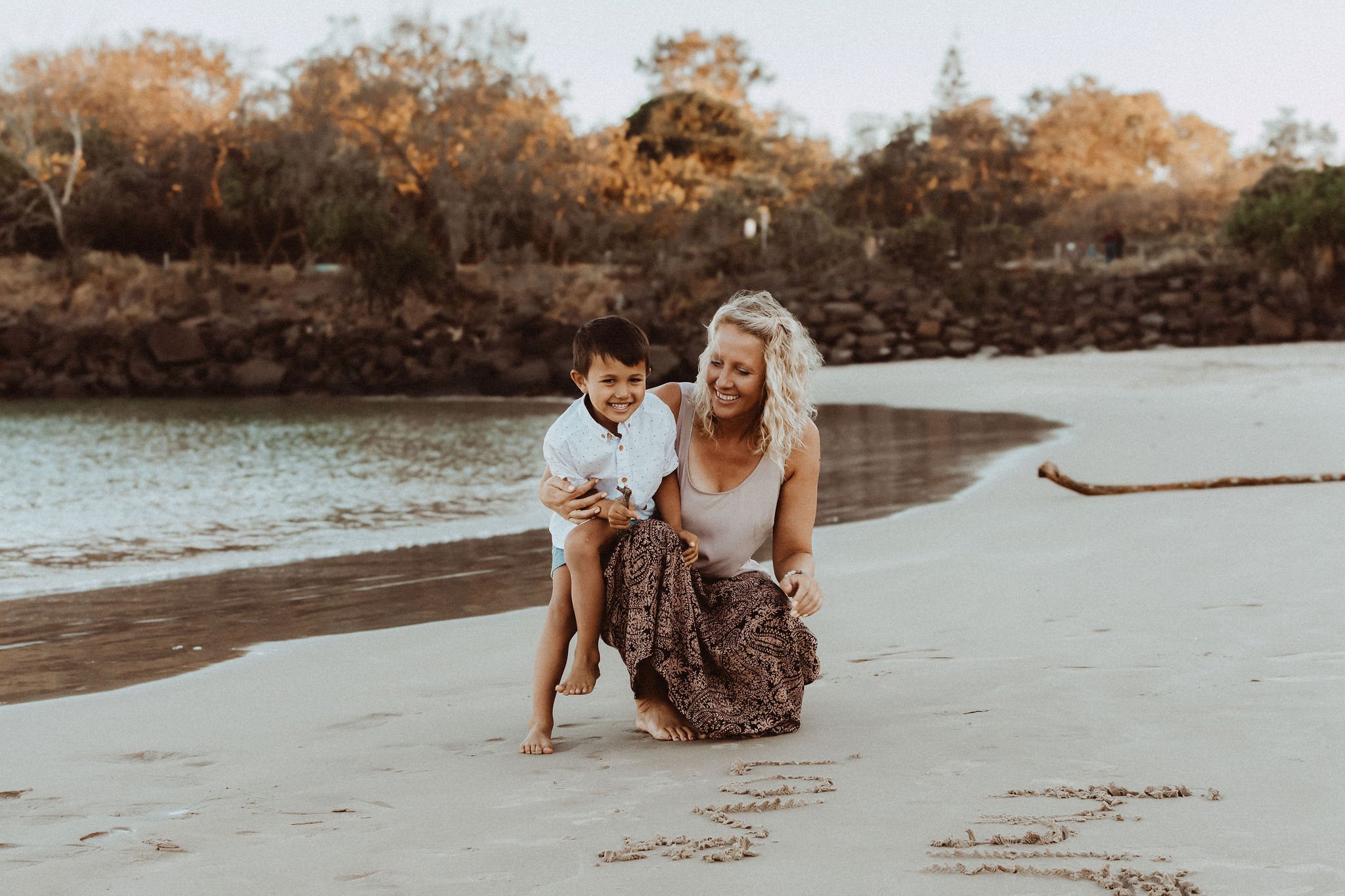 A woman and son smiling on a beach at sunset, with 'family' written in the sand.