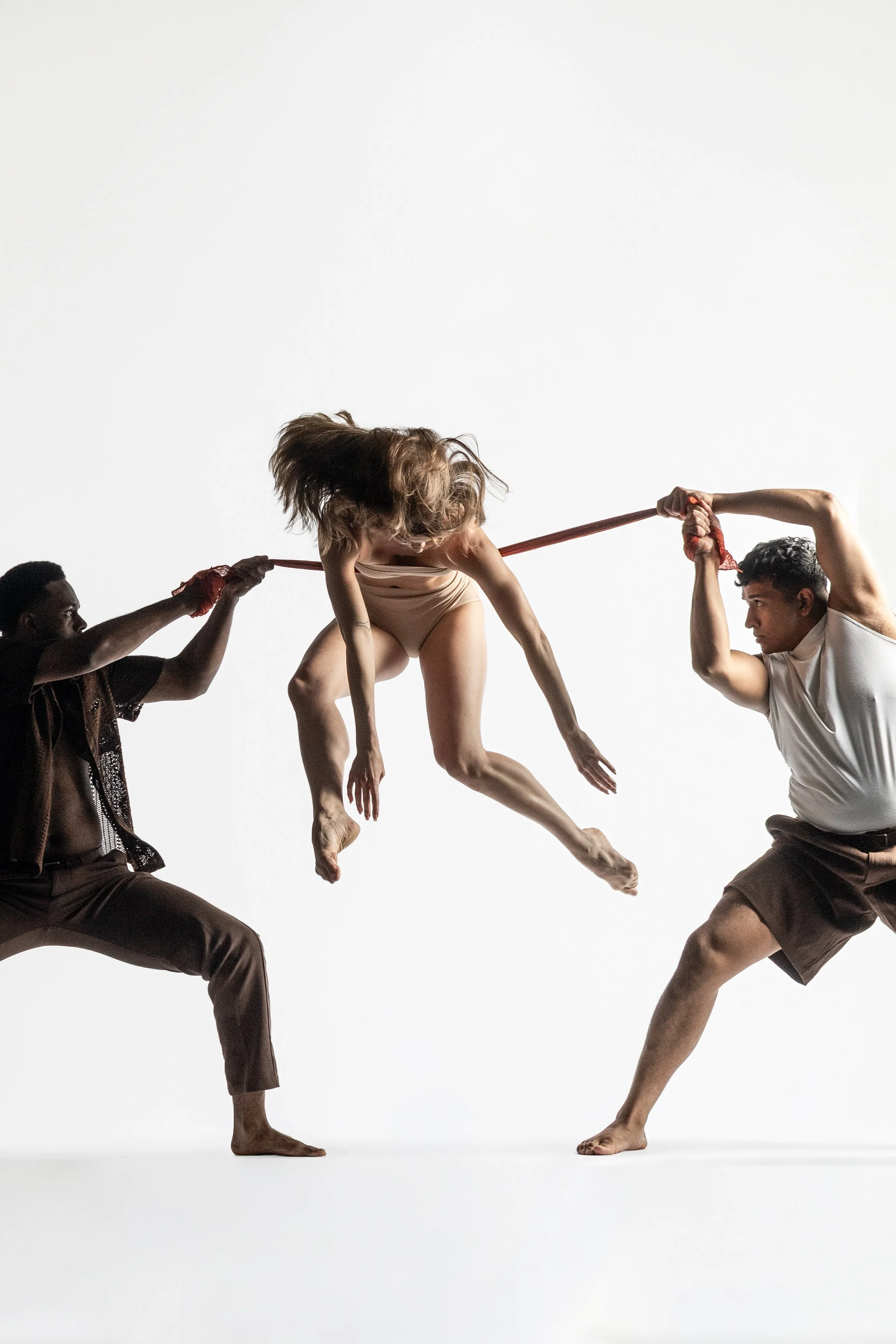 Two men support a woman as she leans sideways and extends her arms and legs in a dance pose against a plain white background.