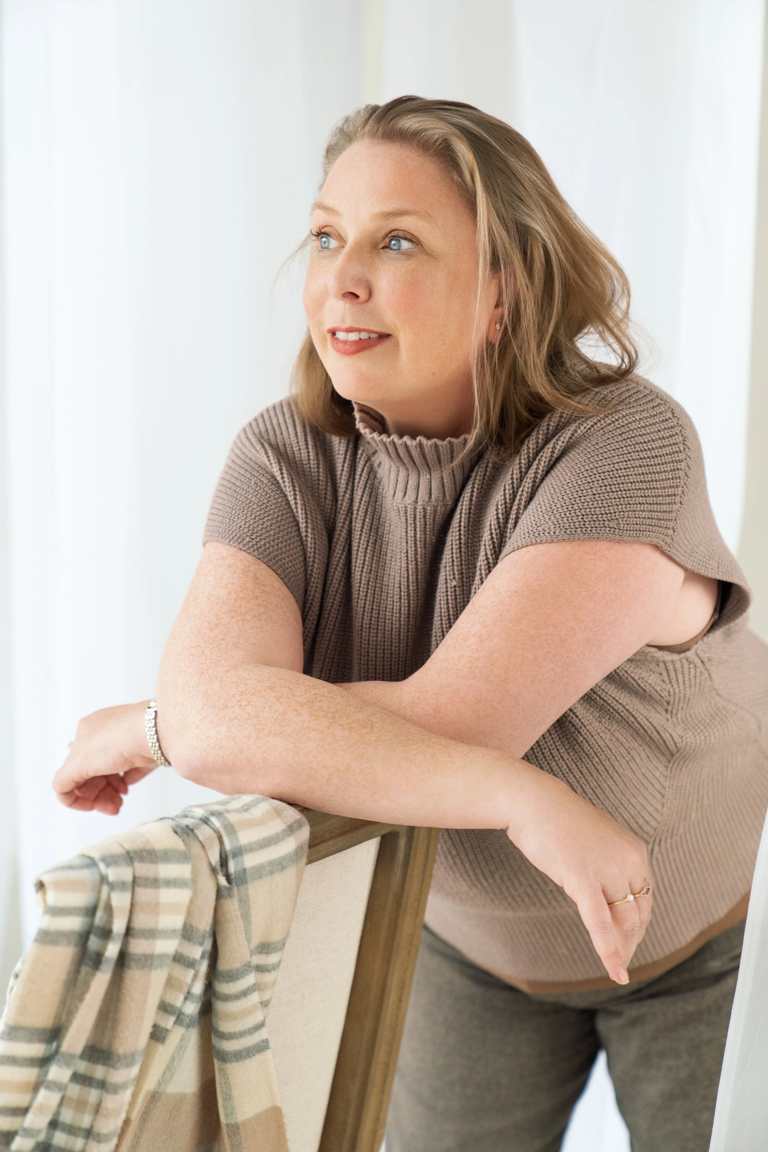 A woman with light brown hair wearing a taupe short sleeve sweater leaning on a chair with a checkered blanket draped over it, looking to the side with a slight smile.