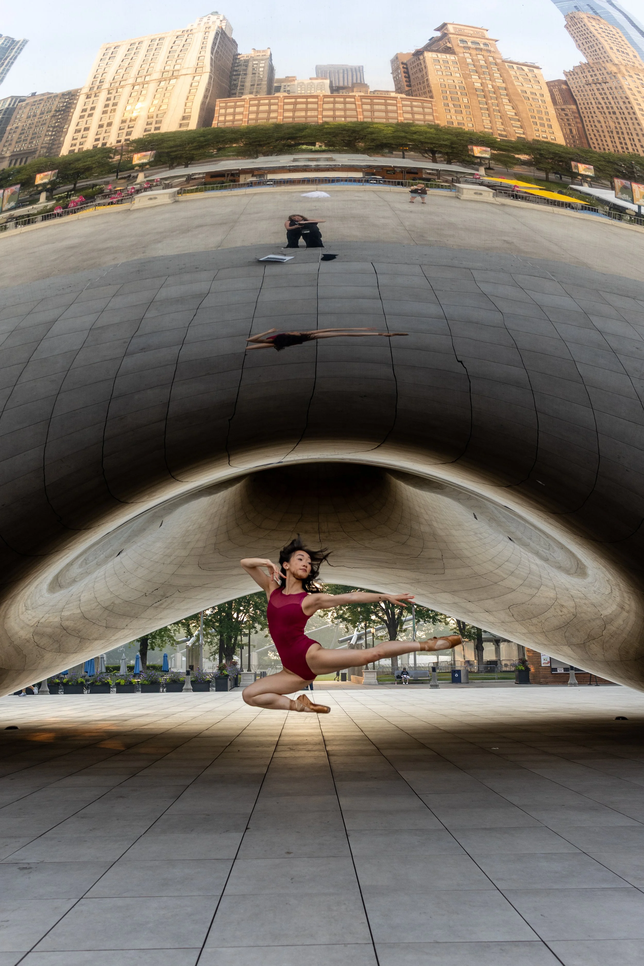 A dancer in a maroon leotard performing a leap beneath the Cloud Gate sculpture, also known as 'The Bean,' in Millennium Park, Chicago, with a city skyline reflected on the sculpture.