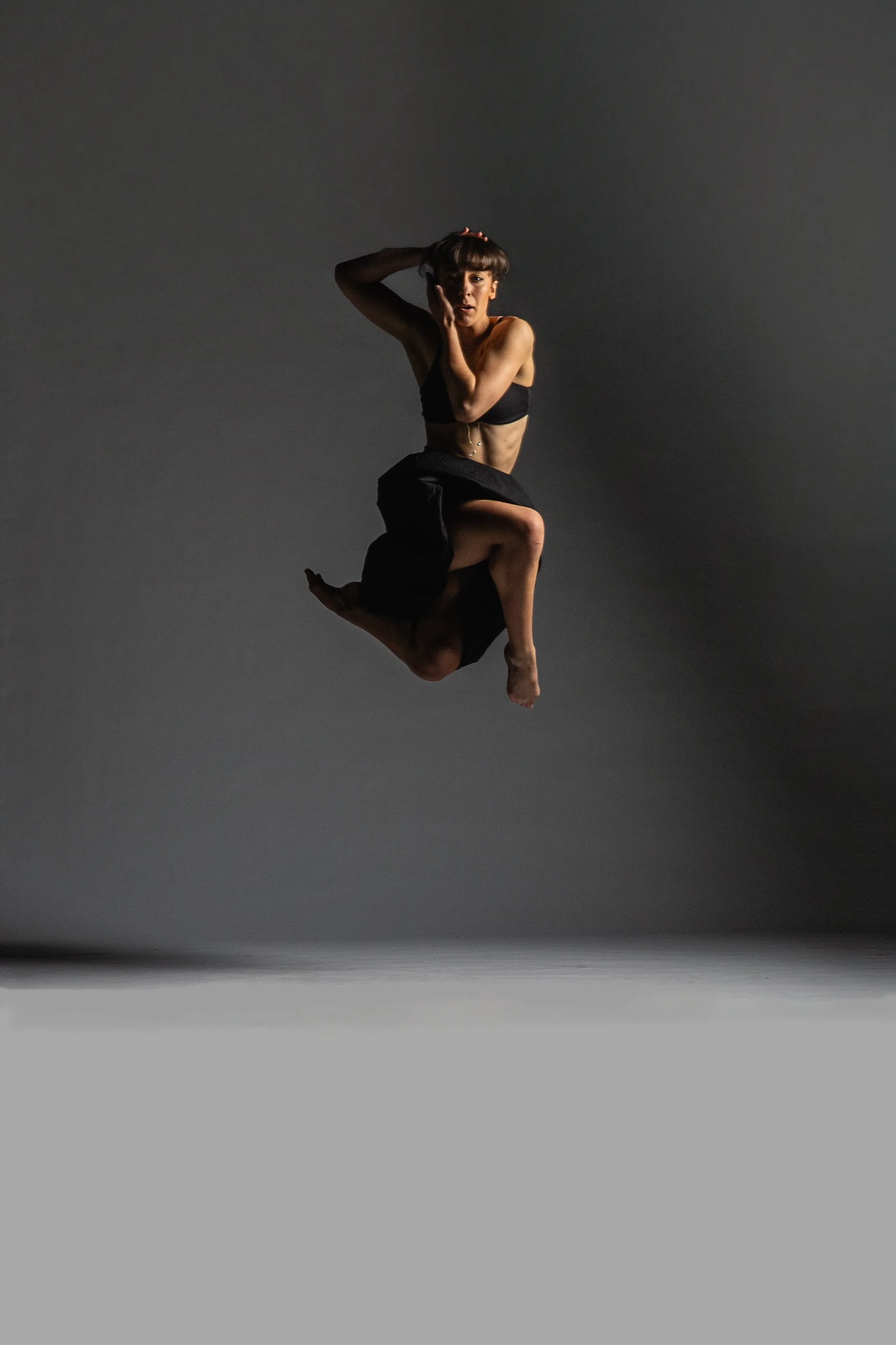 A woman in black sportswear is mid-air performing a dance or acrobatic move against a plain gray background.