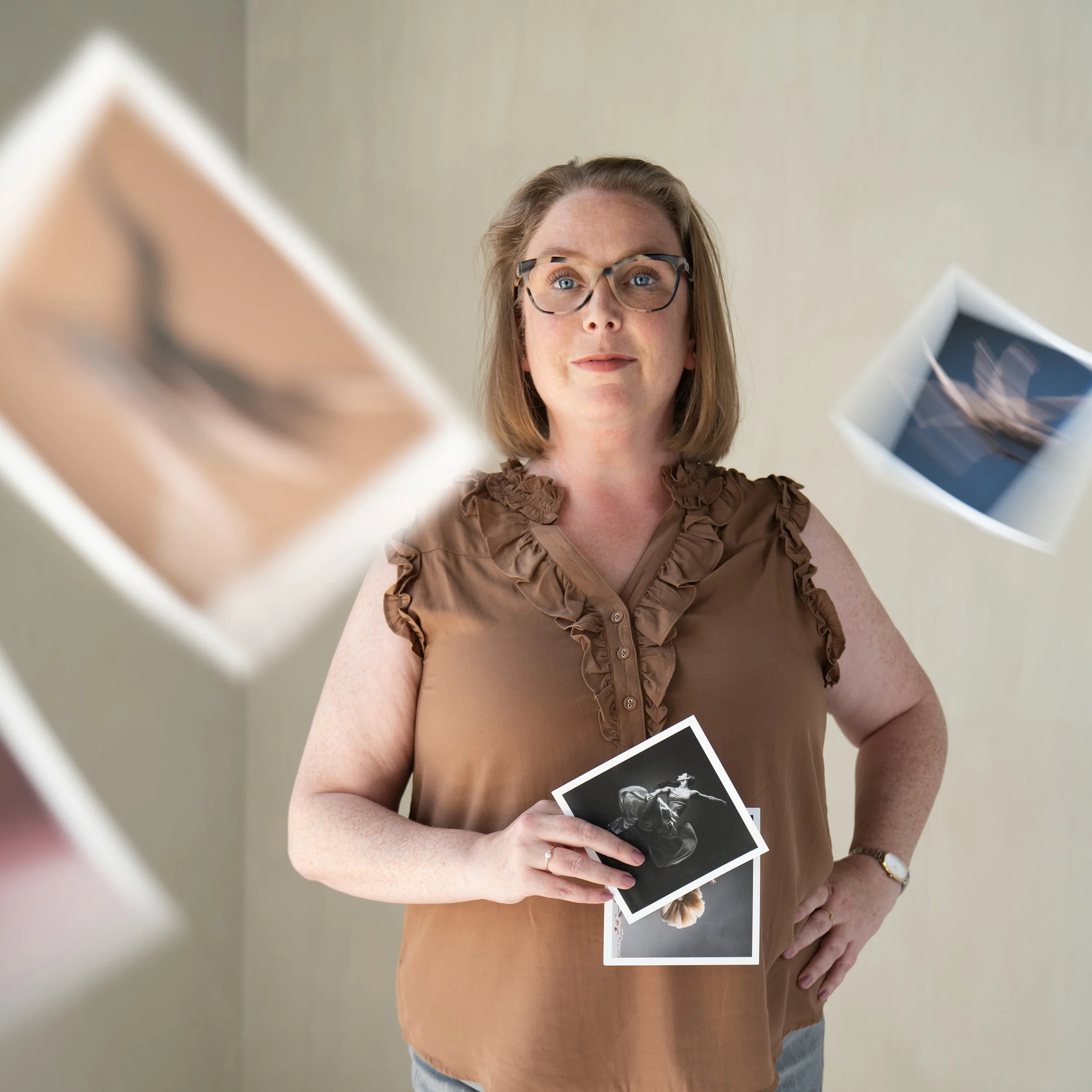 A woman with glasses wearing a brown ruffled blouse holding black and white photos in front of a beige background, with four printed photographs floating around her.
