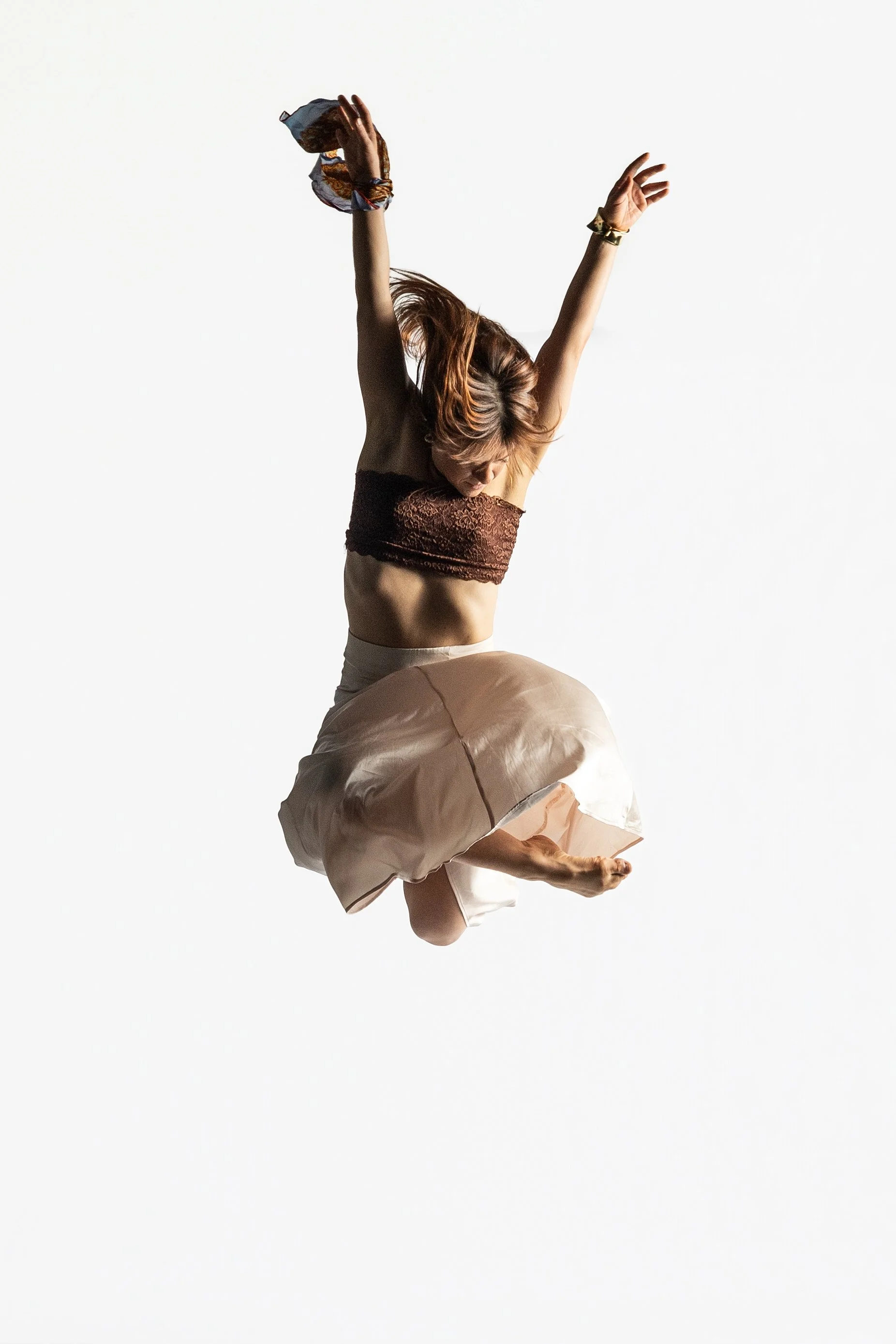 A woman with red hair is captured mid-jump, with arms raised above her head and legs bent, wearing a brown lace top and a flowing beige skirt, against a plain white background.