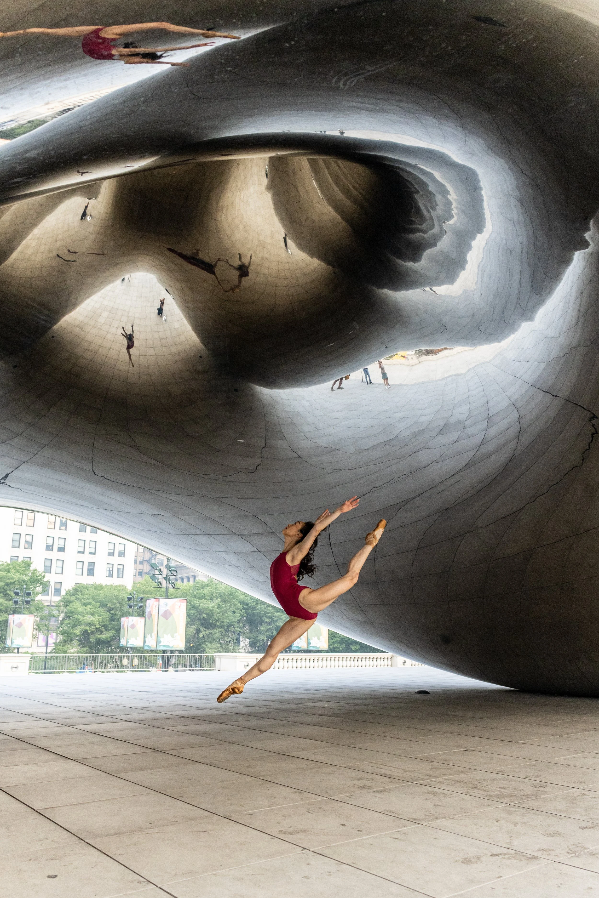 A ballet dancer in a red leotard performing a split leap in front of a large reflective metallic sculpture with a tunnel-like interior. Several people are visible as reflections and in the background outside.