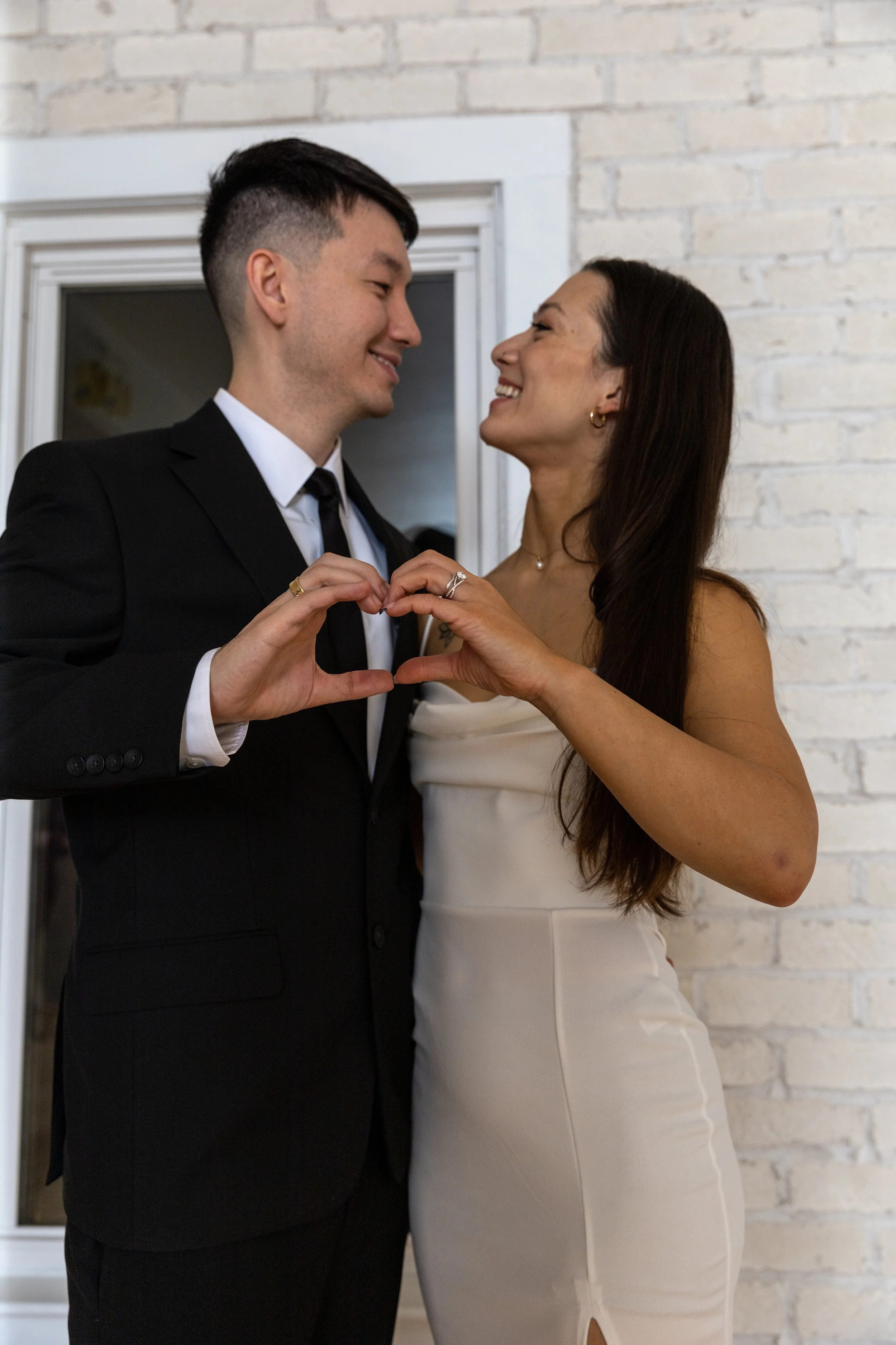 A couple holding hands and making a heart shape with their fingers, smiling at each other, dressed in formal attire, in front of a brick wall.