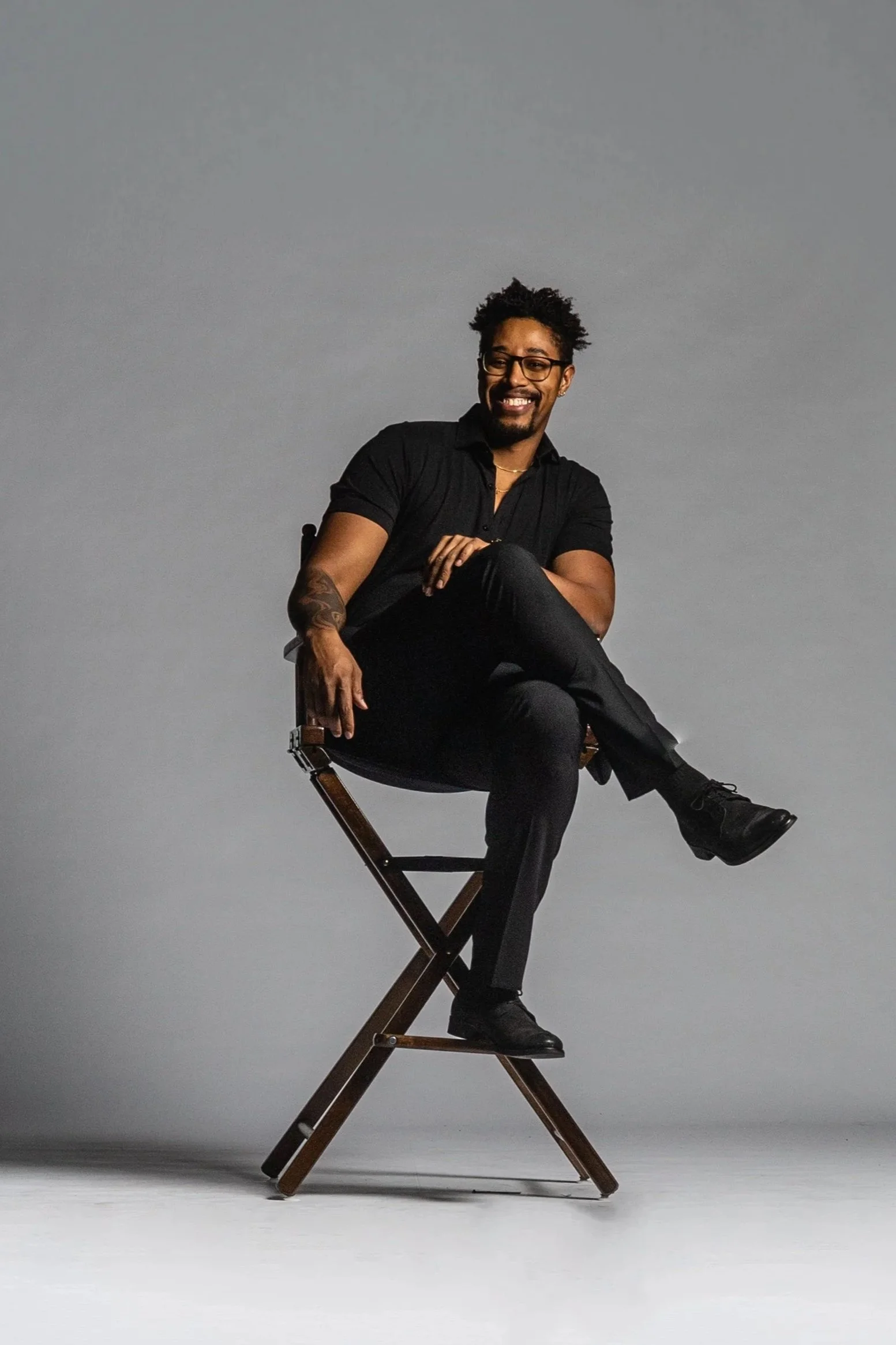 A man with glasses, a beard, and tattoos on his arm, smiling, seated sideways on a director's chair against a plain gray background. He wears a black shirt and black pants, with one leg crossed over the other and his right arm resting on his knee.