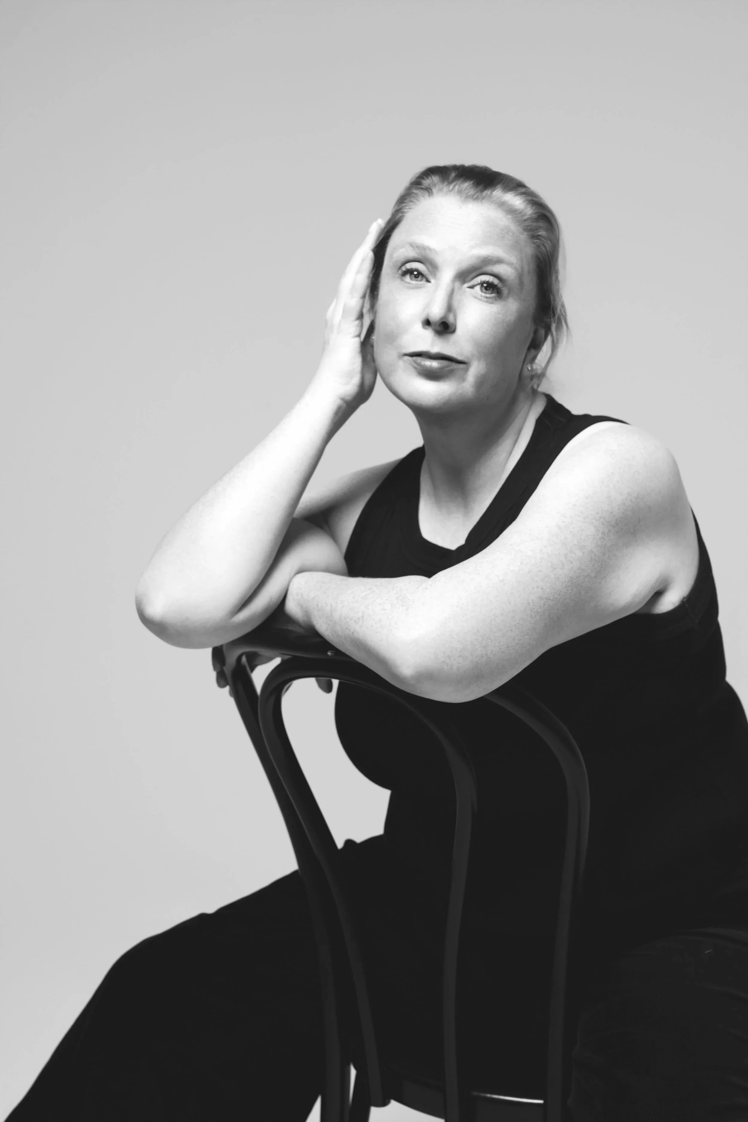 Black and white portrait of a woman with straightforward hair, sitting on a chair, resting her arms on the back of the chair, and touching her head with her left hand.