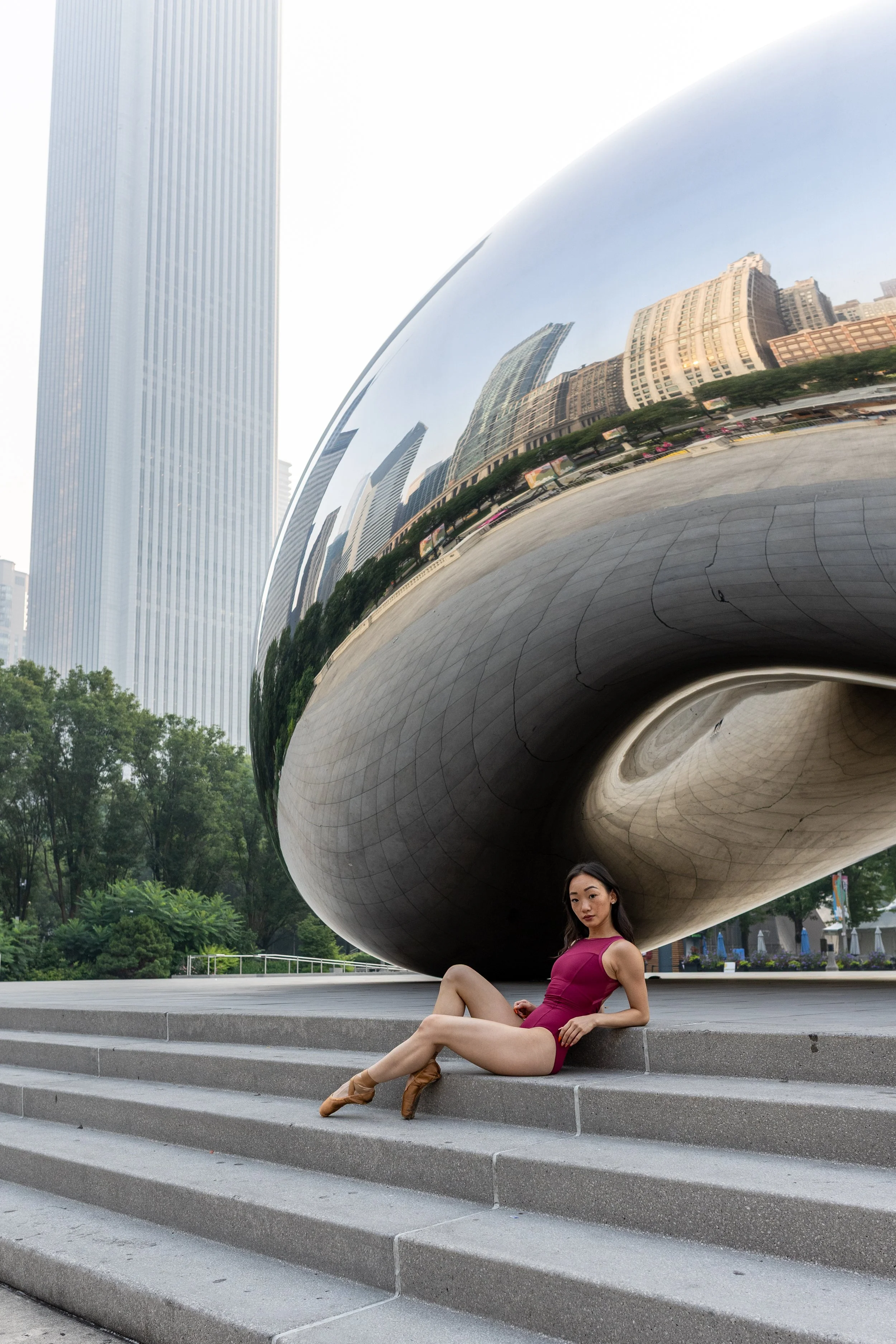 A woman in a purple dress and tan ballet shoes sitting on steps in front of a large reflective sculpture in an urban park setting with tall buildings in the reflection.
