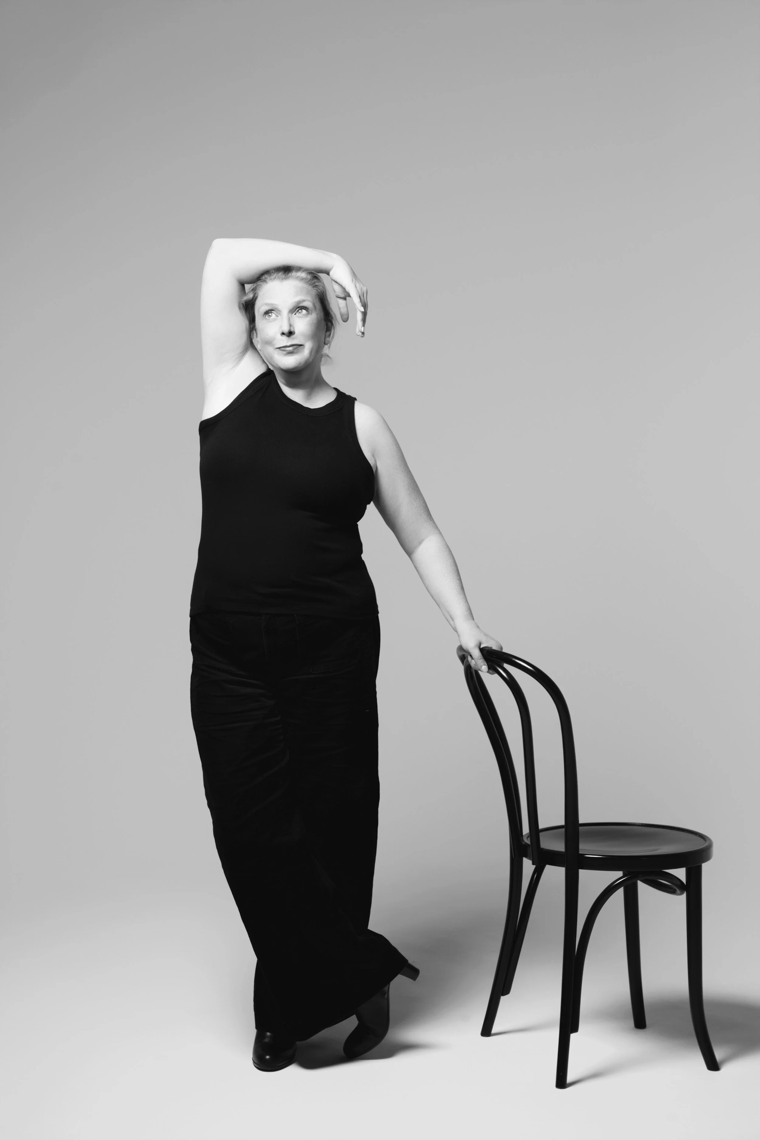 Black and white photo of a woman in a sleeveless top and wide pants standing beside a chair, holding the chair's backrest with her right hand, with her left arm raised and her hand resting behind her head, looking slightly to her left.