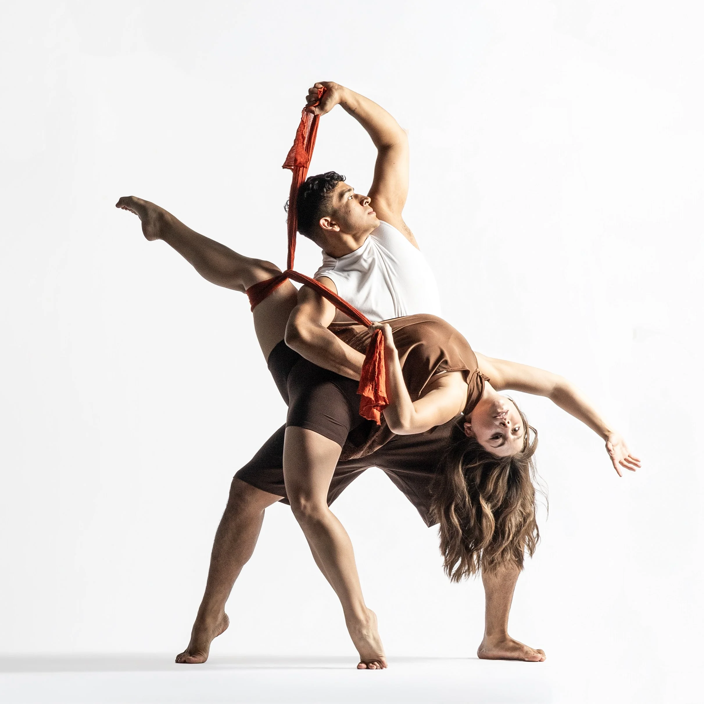 Two women practicing acro yoga, one supporting the other in a handstand while holding a red fabric, against a plain white background.