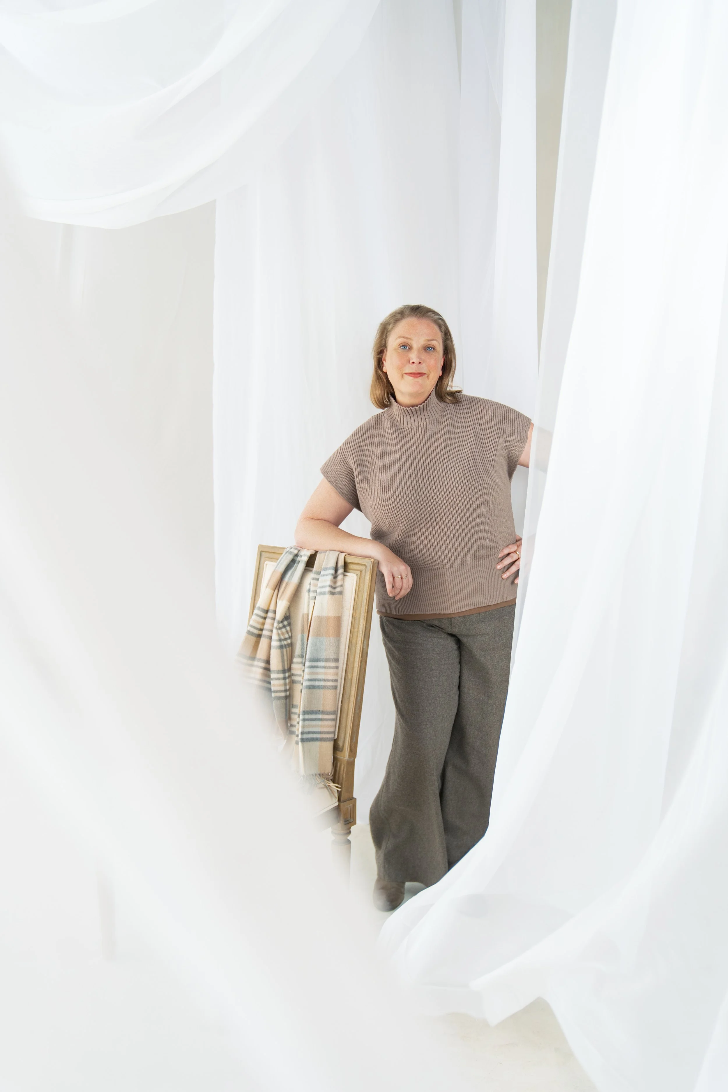 A woman standing among white curtains, wearing a beige sleeveless sweater and gray pants, with one hand on her hip and the other resting on a wooden chair draped with a plaid scarf.