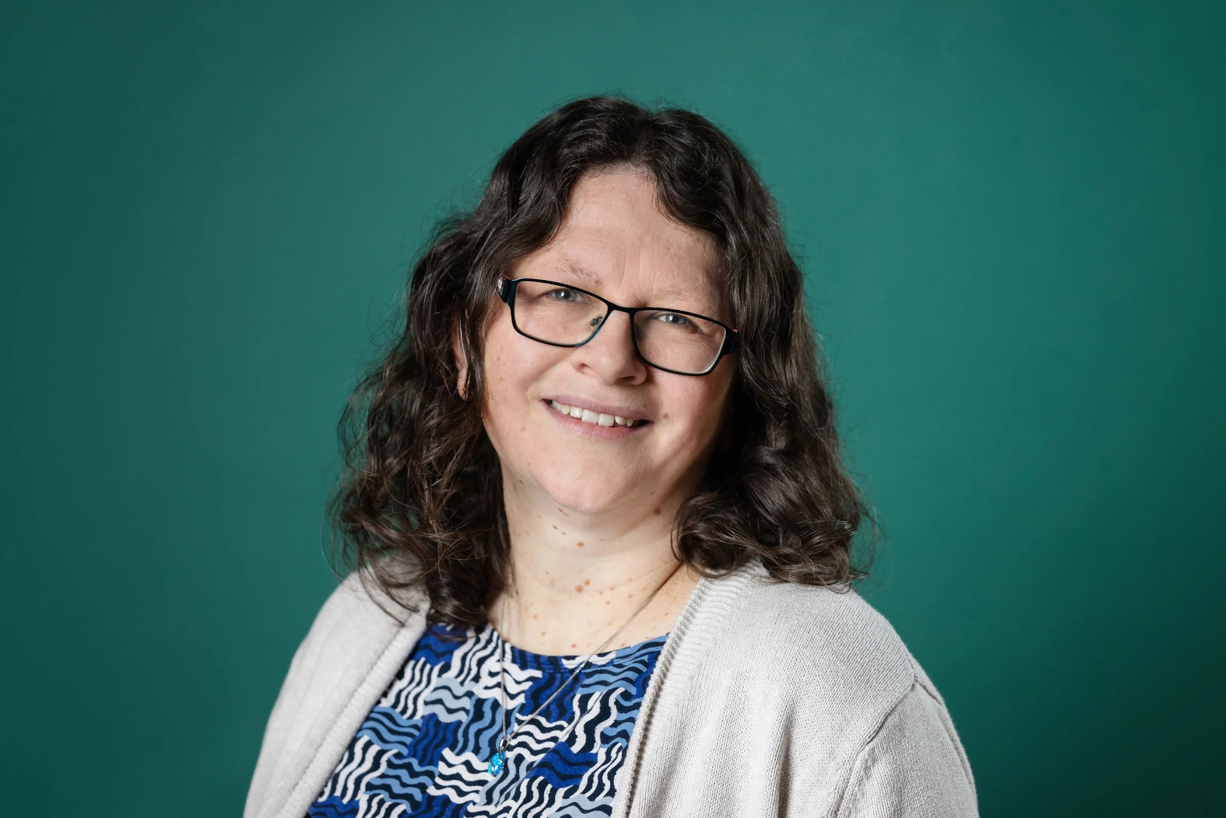 Portrait of a woman with curly brown hair, wearing glasses, smiling, dressed in a blue patterned top and light-colored cardigan, against a teal background.