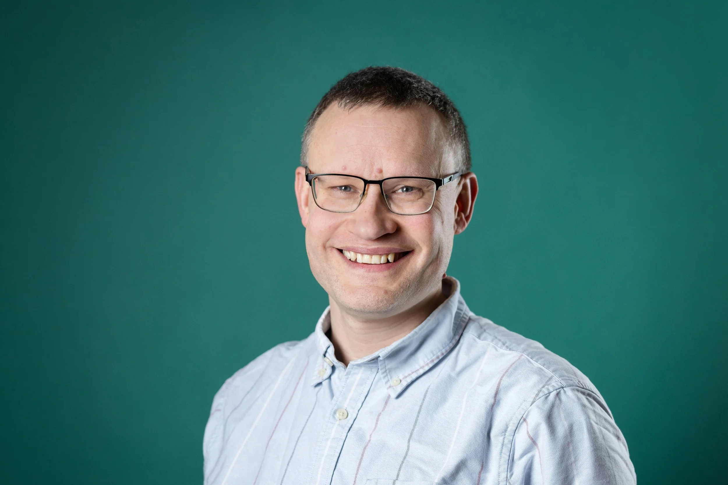 A smiling man with short brown hair, glasses, and light skin, wearing a white button-up shirt with thin vertical stripes, posed against a solid green background.