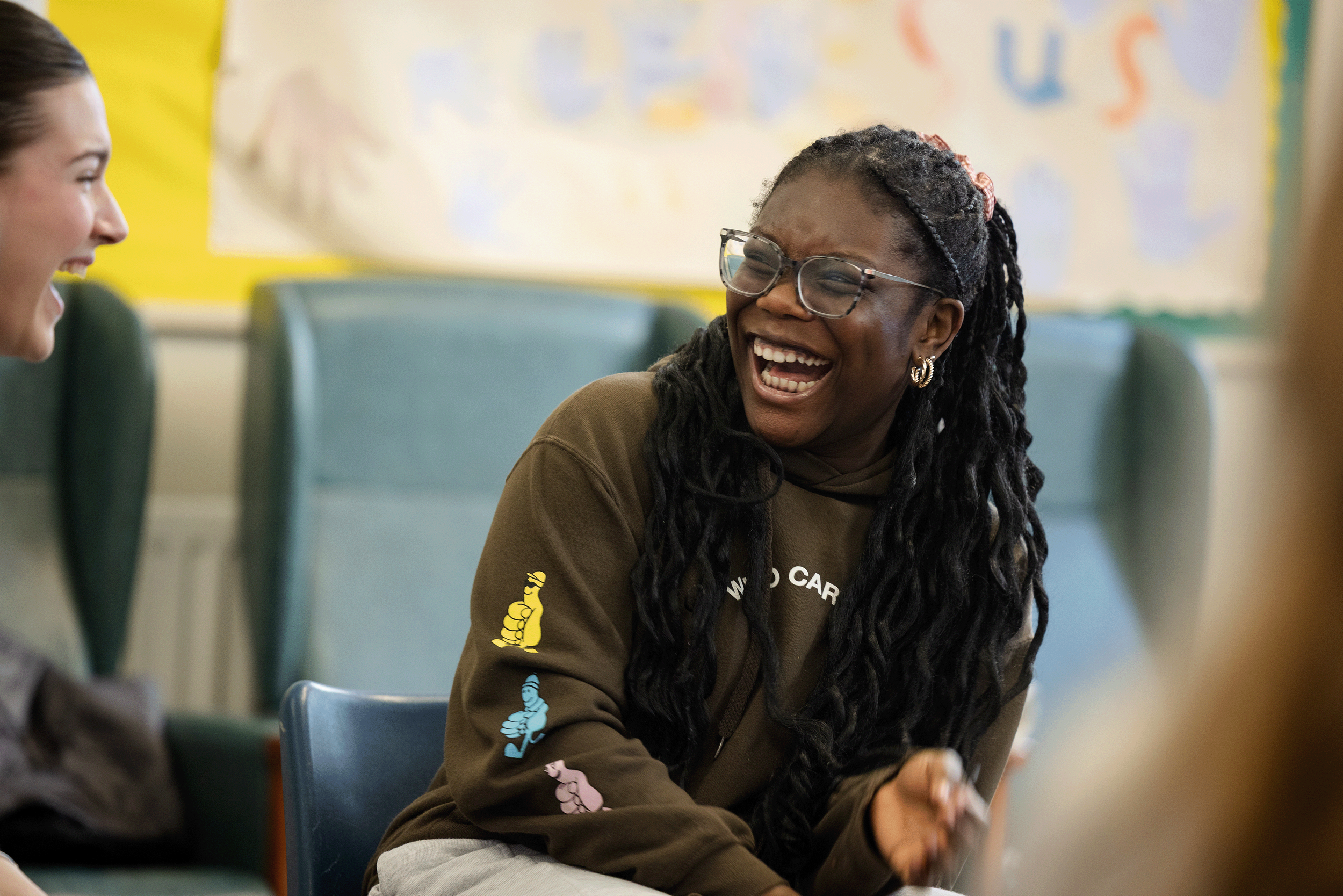 Two girls laughing and talking in a classroom or community space, with a yellow and colourful wall in the background.