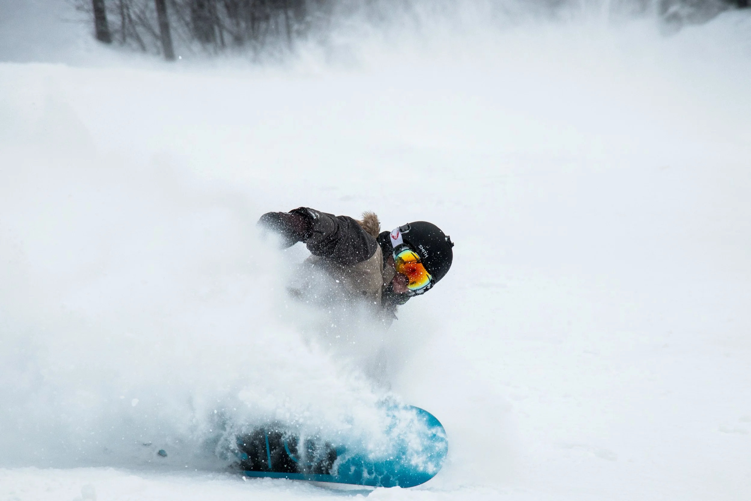 A snowboarder wearing a black helmet, goggles, and a dark jacket turns sharply into powder snow, sending a wave of snow flowing out from their board.