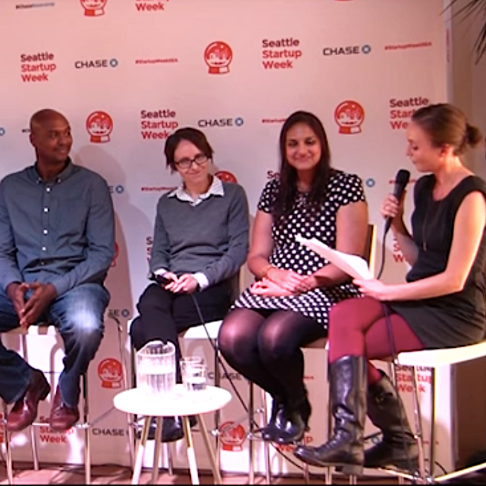 Four people with diverse genders and skin tones wearing professional clothing are seated on a stage. The person on the right speaks into a microphone while the others look towards them.