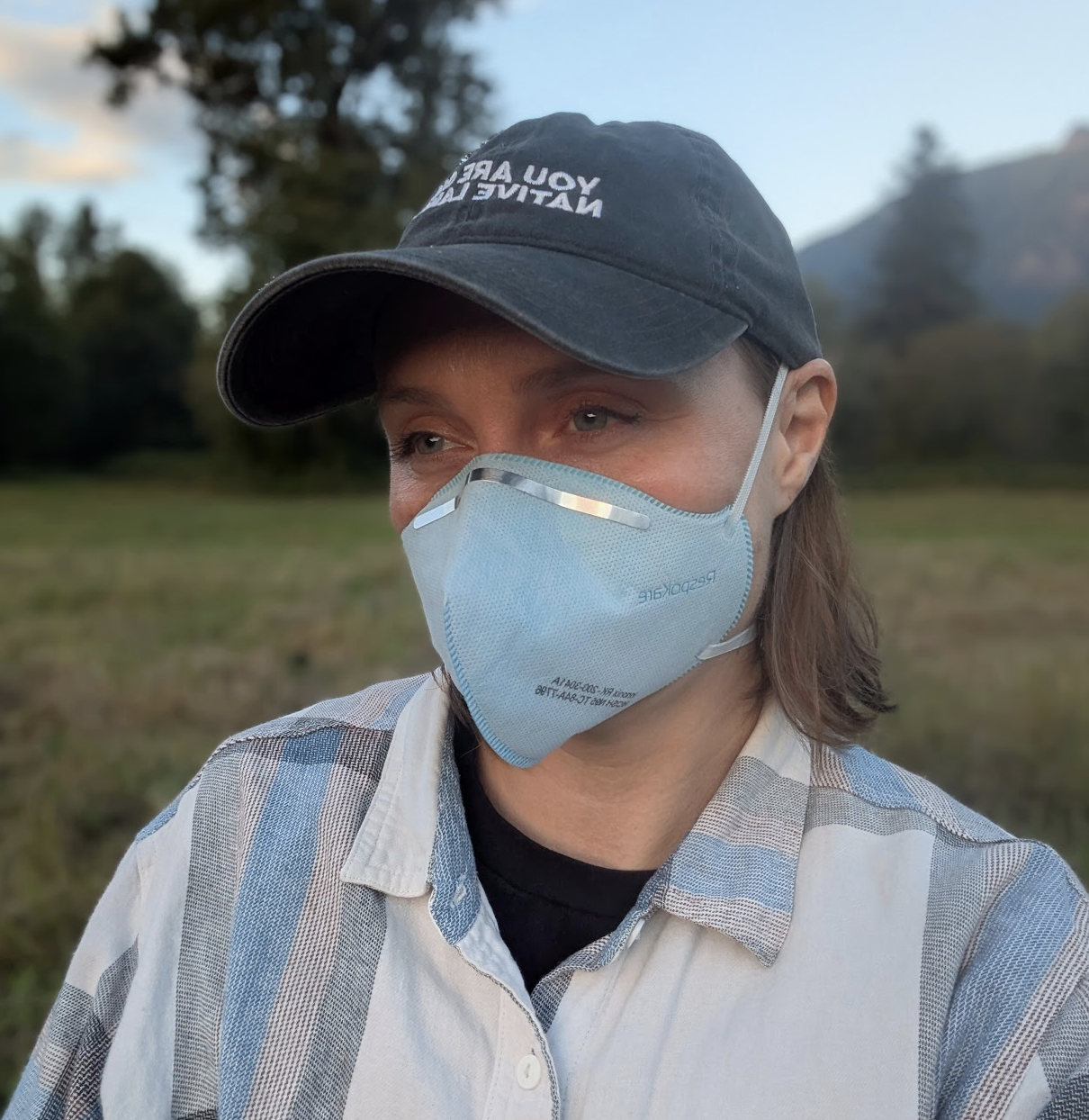 M Burwell, A white person with blue eyes and brown hair, wears a blue respirator mask and a cap that reads "you are on native land" while in a meadow
