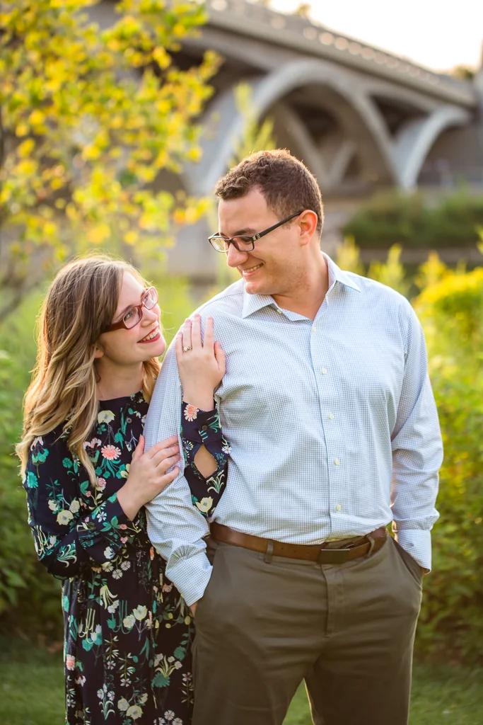 Woman poses standing behind man and snuggling his arm while featuring her engagement ring. Couple smiles at each other in front of bridge and sunset at the Scioto Mile Riverfront in Columbus OH during their engagement session.