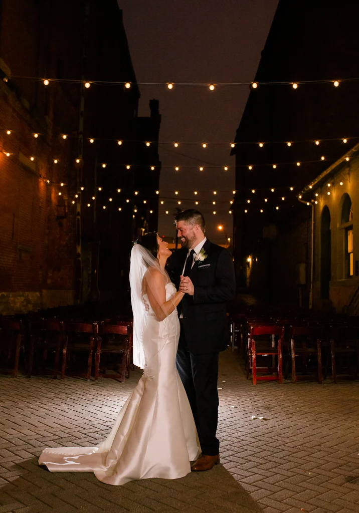 Night portrait of Bride and groom dancing under twinkling string lights in brick alleyway after their wedding at Via Vecchia.