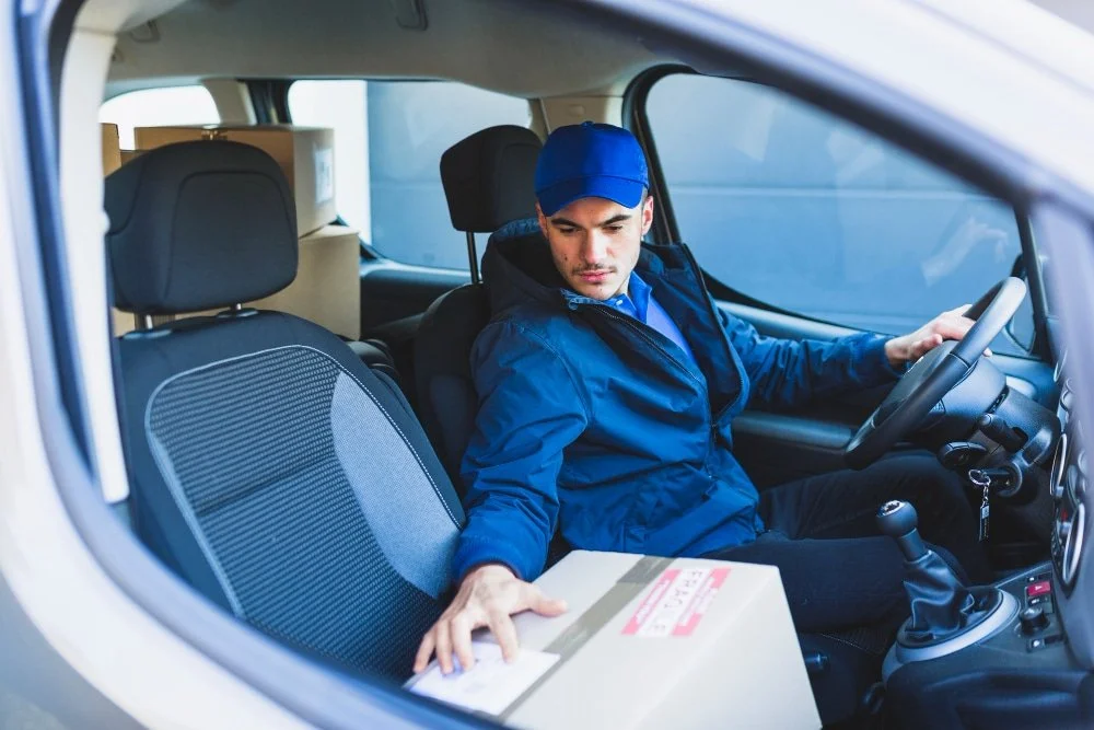 A delivery driver sitting in a vehicle with a box on his lap, wearing a blue cap and jacket, looking at the box.
