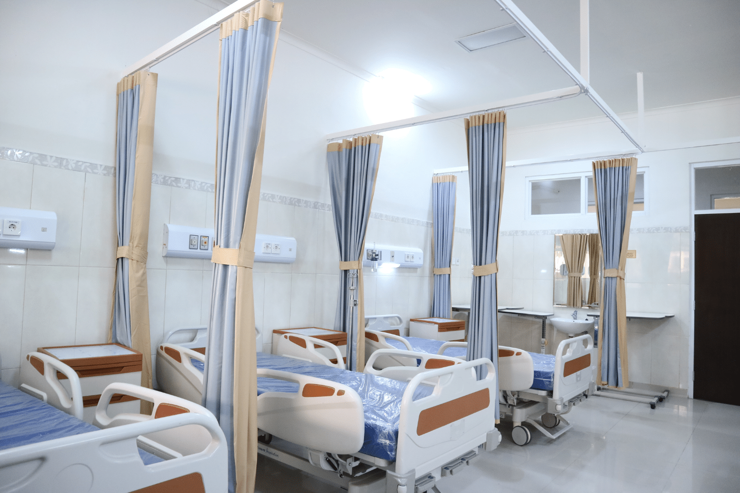 Hospital ward with multiple beds separated by curtains, with medical equipment and a sink in the background.