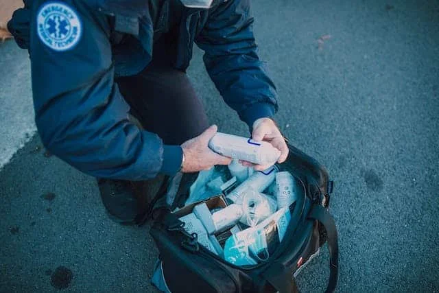 Person in a dark uniform kneeling on the ground, holding a smartphone, with an open medical bag containing supplies nearby.