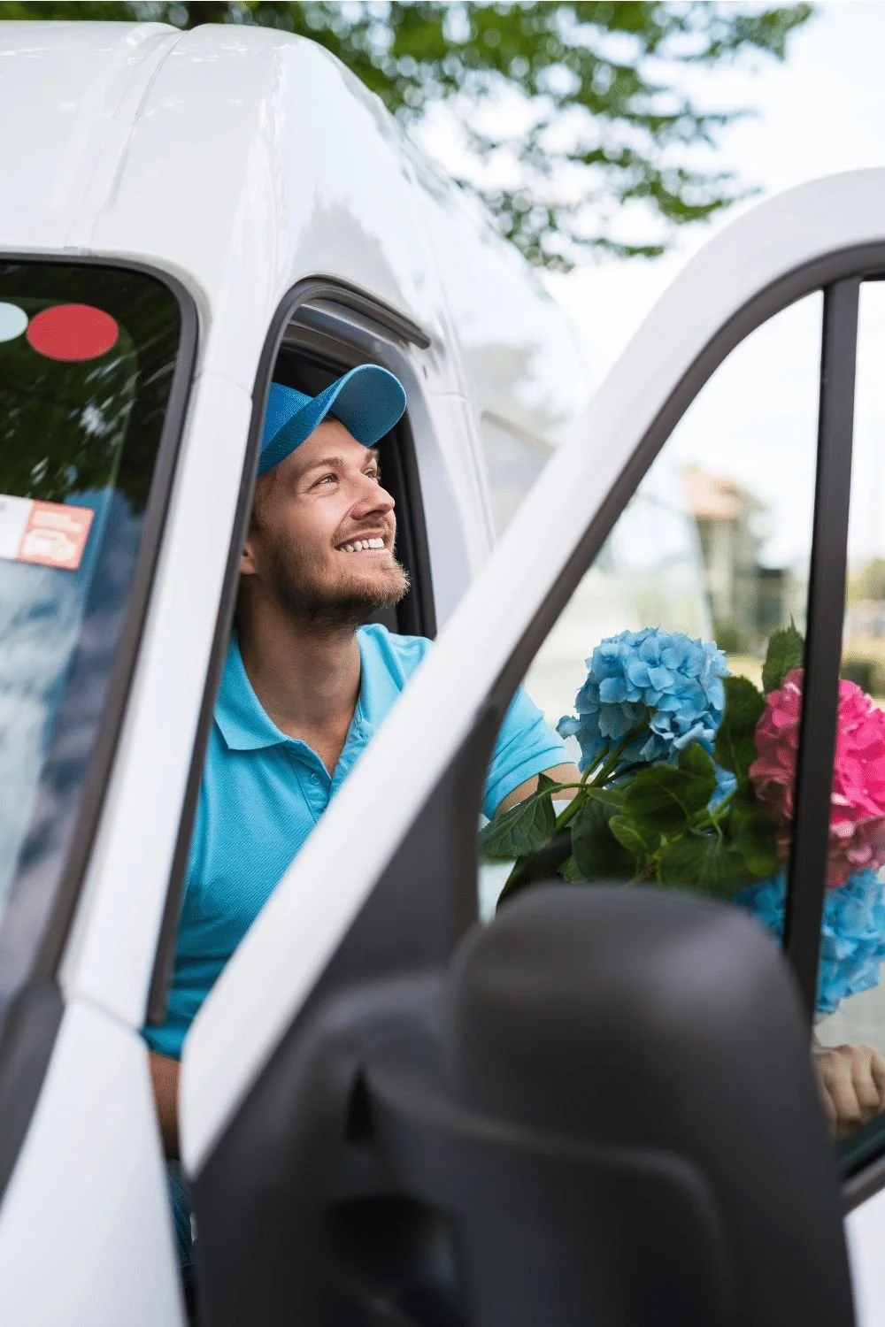 A man wearing a blue cap and shirt smiling while sitting inside a delivery van with flowers.