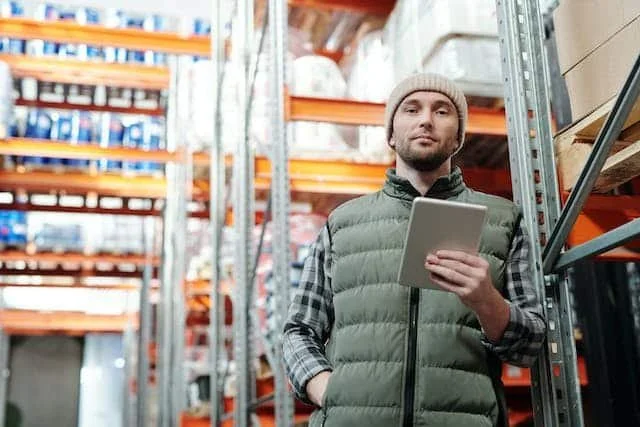 A man standing in a warehouse aisle holding a tablet, surrounded by metal shelving with boxes and supplies.