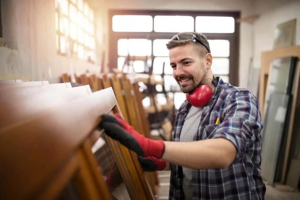 A smiling man in a plaid shirt and safety glasses is working in a woodworking workshop, sanding or inspecting a wooden piece.
