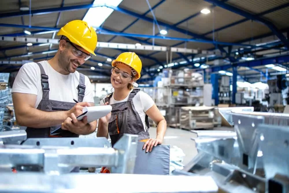 Two factory workers, a man and a woman, wearing yellow safety helmets and safety glasses, are smiling and looking at a clipboard in an industrial warehouse.