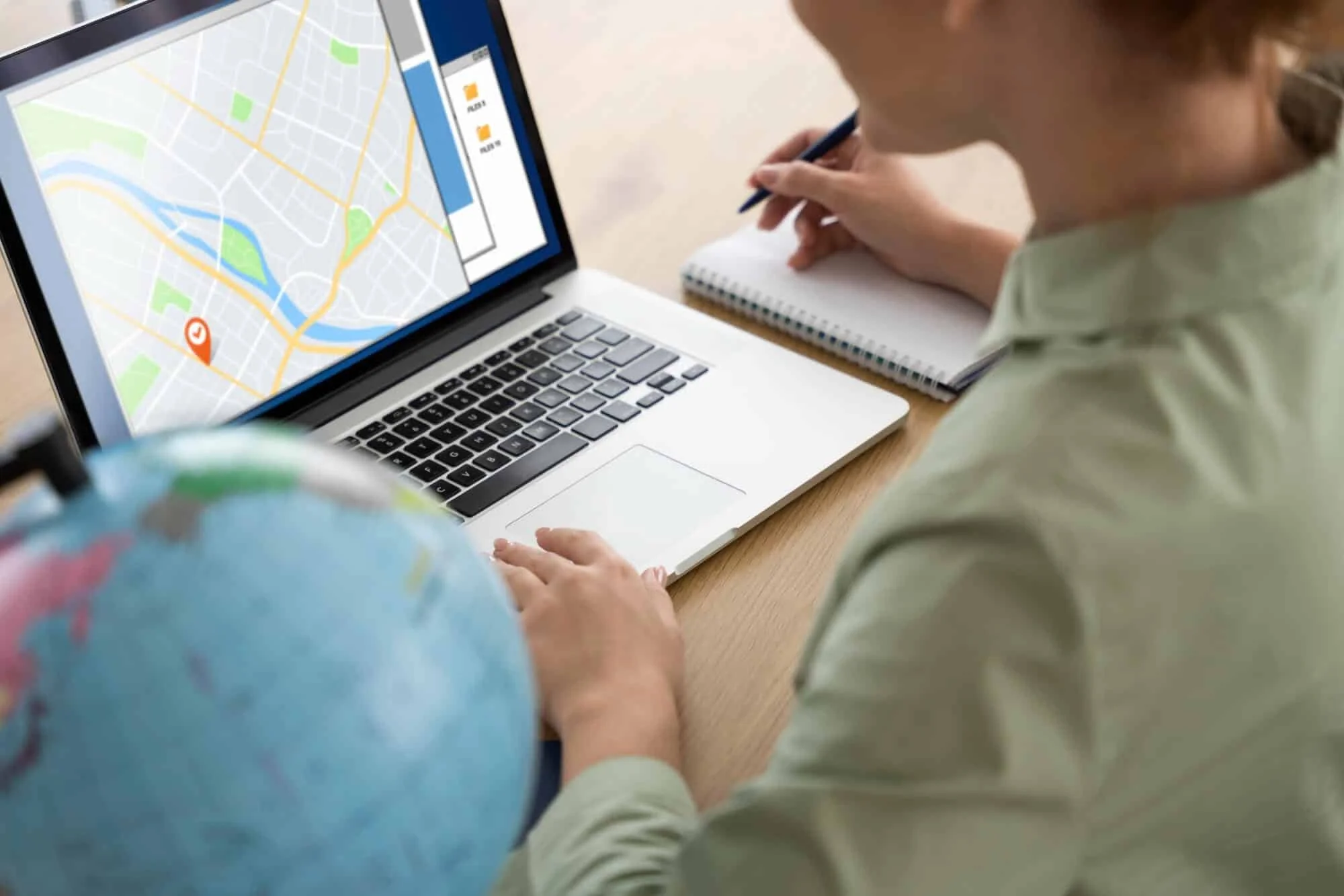 A person using a laptop with a map and navigation app open on the screen, taking notes in a notebook on a wooden desk.