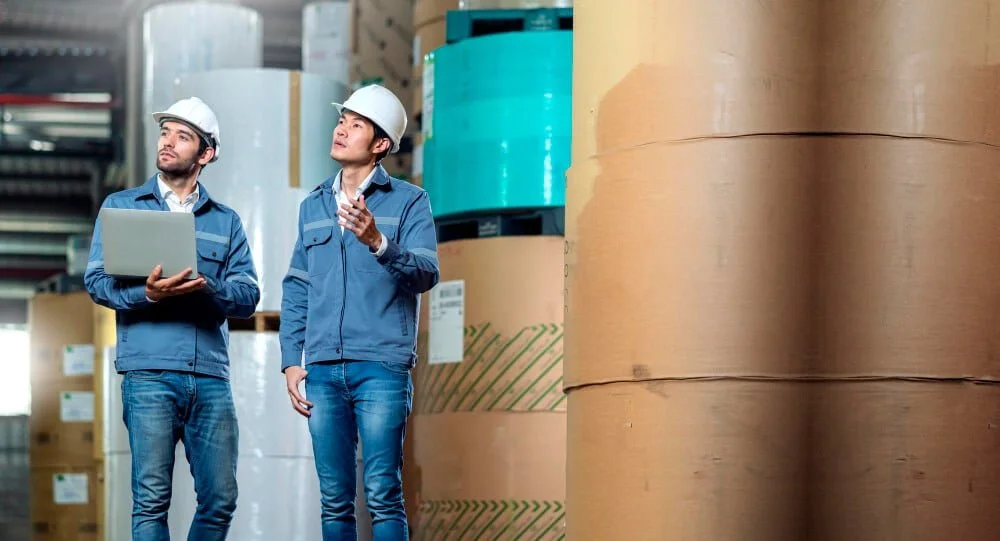 Two men wearing safety helmets in a warehouse, one holding a laptop and the other making a gesture, surrounded by large rolls of paper or material.