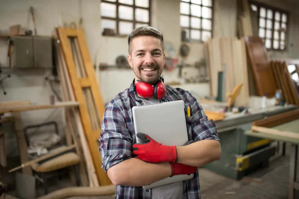A man in a woodworking shop holding a laptop and smiling, wearing red ear protection and red gloves, surrounded by wooden furniture and tools.