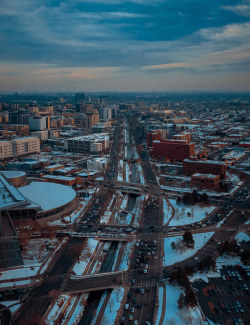 Aerial view of a cityscape with snow-covered roads, buildings, and traffic on a winter day, with a cloudy sky overhead.