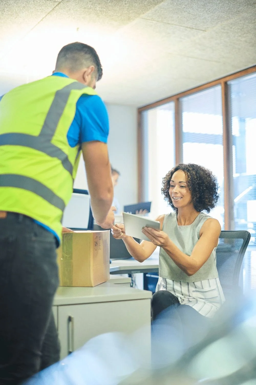 A woman sitting at a desk exchanging a package and a tablet with a man in a yellow safety vest in an office.