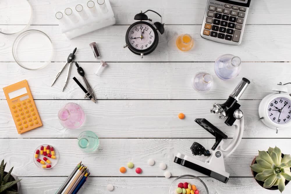 Flat lay of laboratory and office supplies including a clipboard, compass, pills, test tubes with liquids, a microscope, clocks, calculator, and pens on white wooden surface.
